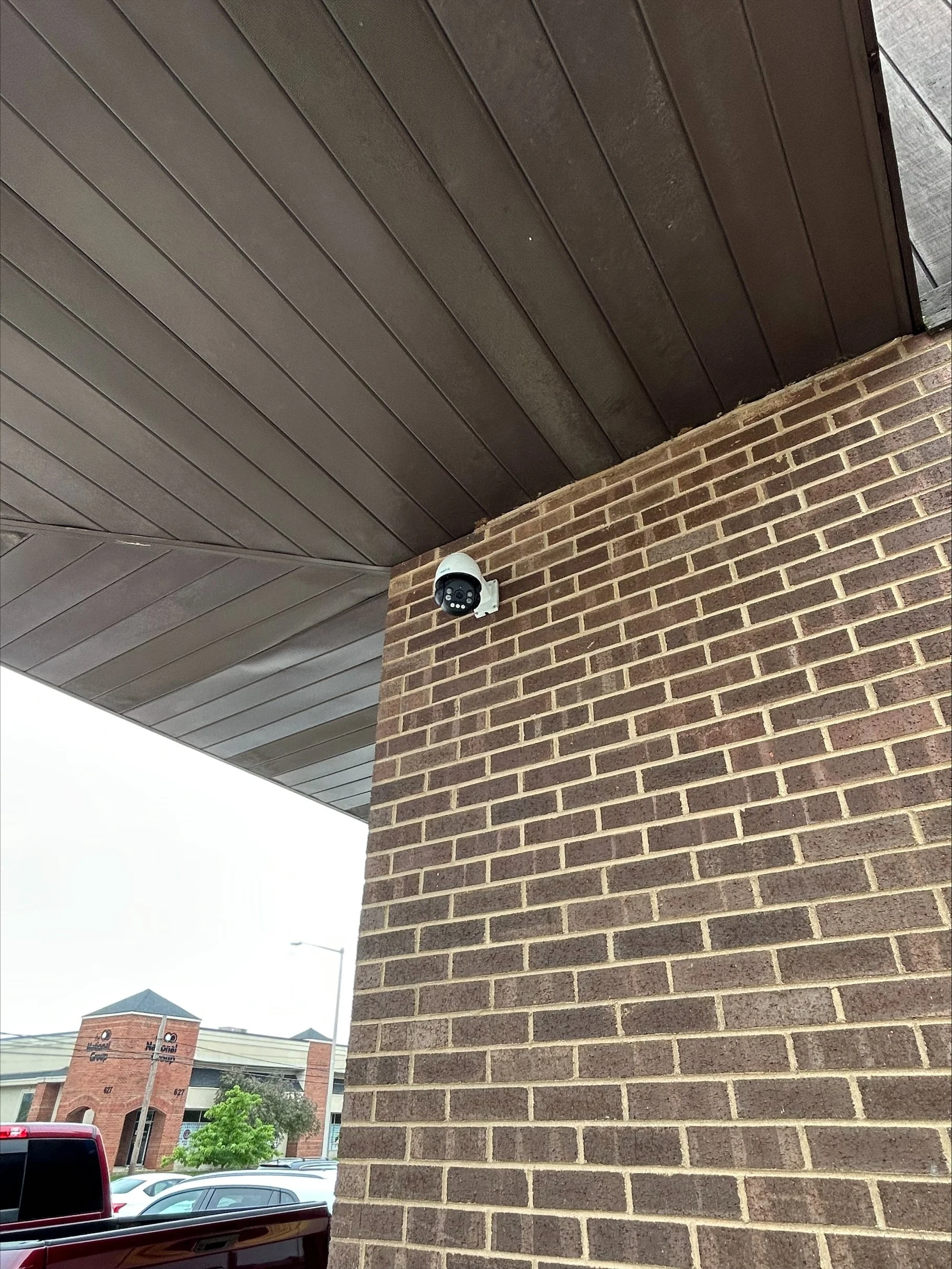Security camera mounted on brick wall under a brown wooden ceiling, with part of a parking lot and building visible in the background.