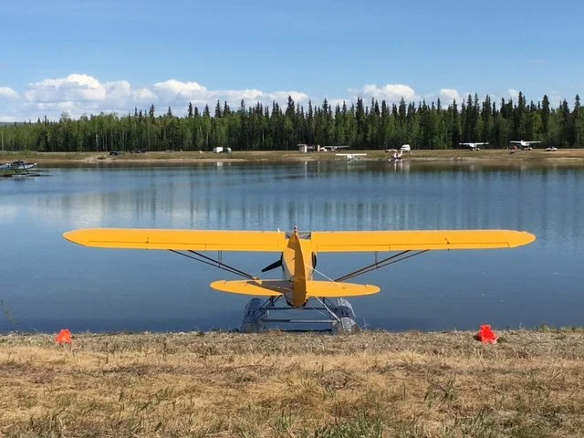 Yellow seaplane on the shore in Fairbanks Alaska with a forested shoreline and small airplanes parked in the background under a partly cloudy sky.