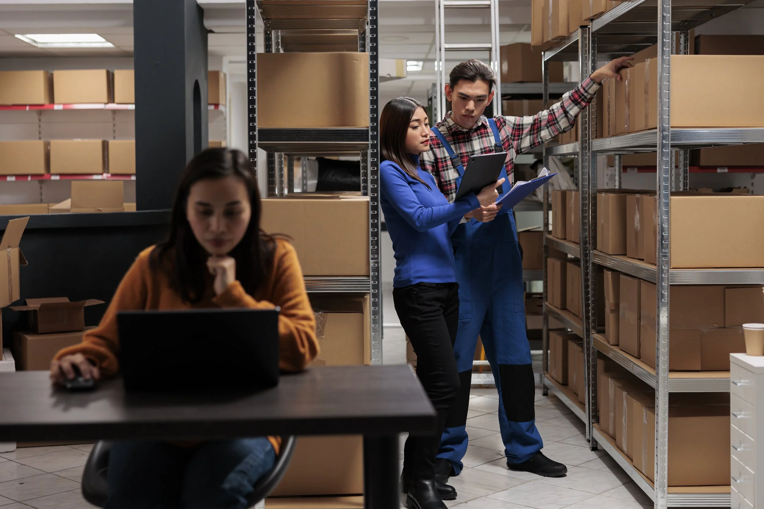 Two warehouse workers, a woman and a man, are examining inventory shelves filled with boxes. The woman is holding a tablet and a clipboard, the man is looking at the shelves while holding a box. A third person, a woman, is sitting at a table using a laptop in the foreground.