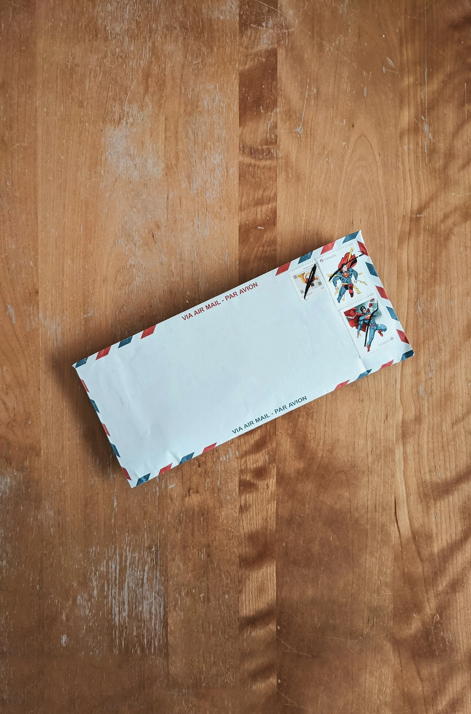 A white envelope with red and blue border stripes, laying on a wooden surface, with a Canadian postage stamp featuring a comic book superhero.
