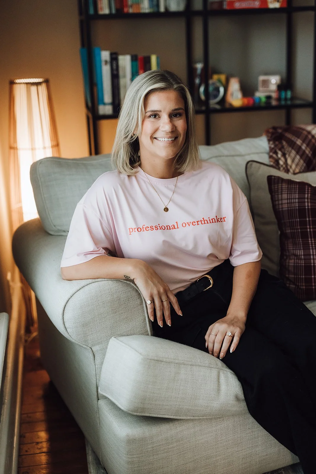 A woman sitting on a beige couch in a living room, smiling at the camera, wearing a pink t-shirt that says "professional overthinker" in red text, with a gold necklace and rings, in front of a bookshelf with books and decorative items.