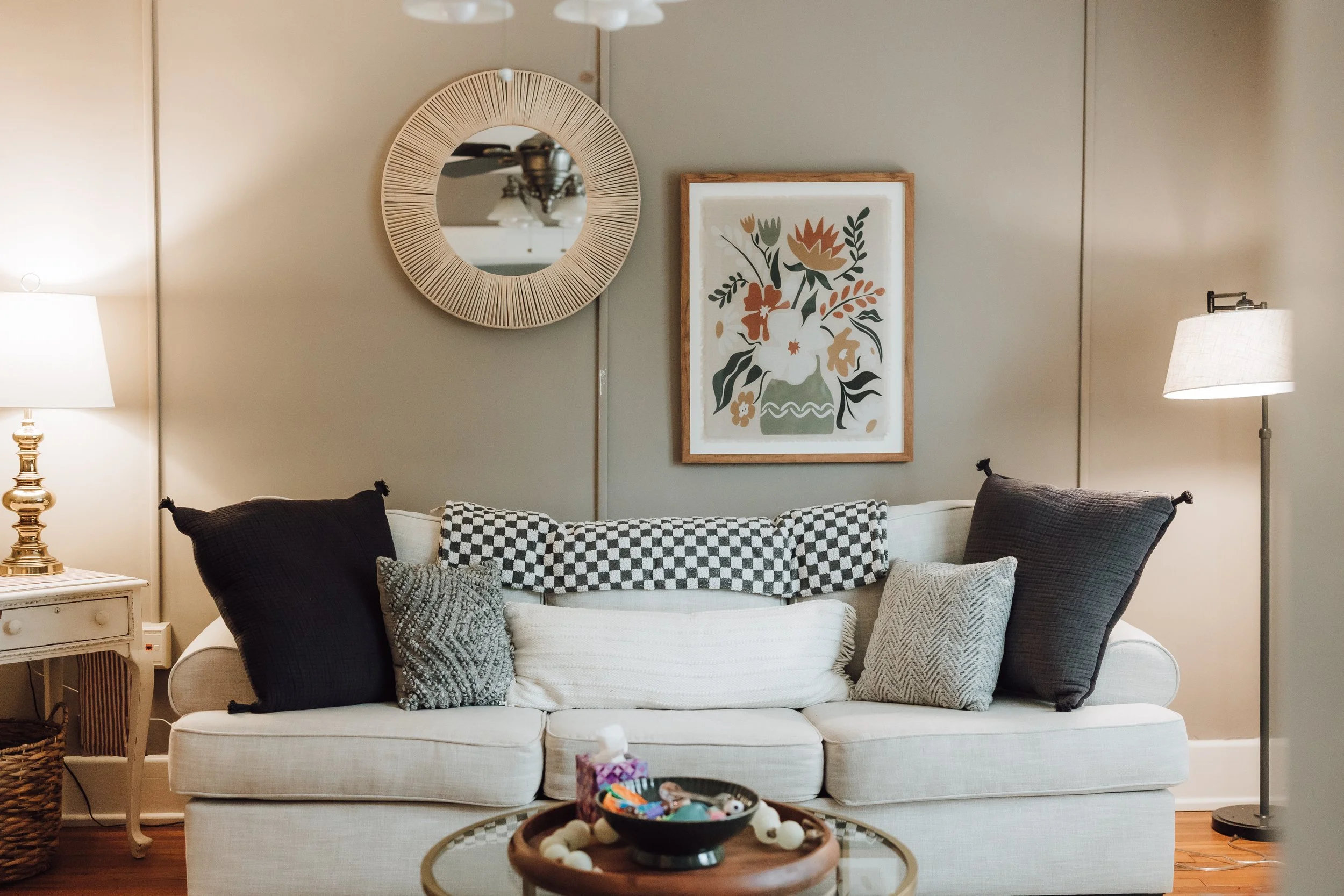 Living room with white sofa adorned with black and white checkered and textured gray pillows. On the wall behind the sofa, there is a round mirror with a woven frame and a framed abstract floral art print. Two table lamps with white shades are on each side of the sofa, and a small wooden side table with a drawer is visible on the left, topped with a gold lamp. A coffee table in front holds a bowl filled with colorful objects, with a basket on the floor beside the side table.
