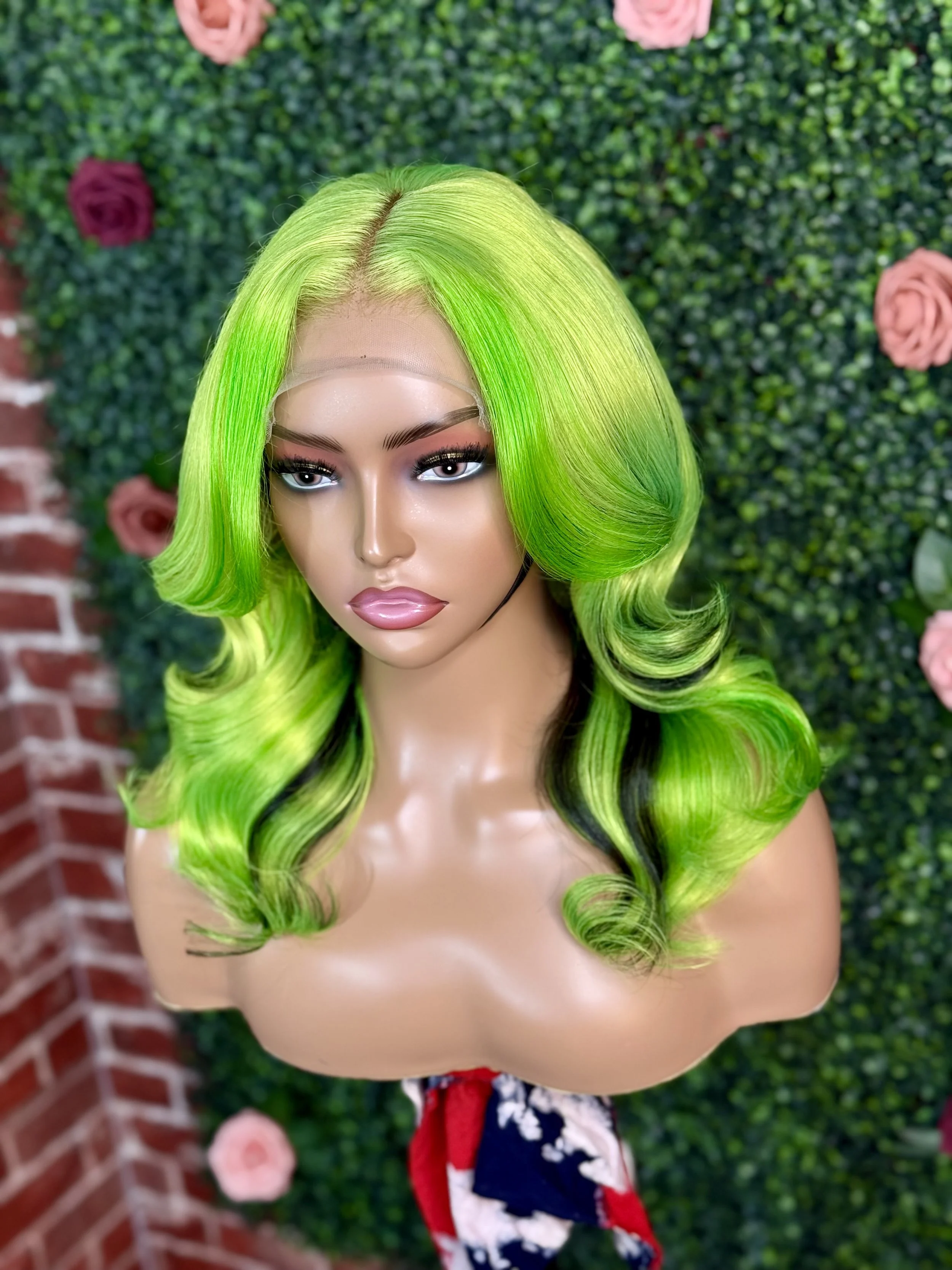 A mannequin with bright green hair styled in loose waves stands in front of a green hedge decorated with pink roses.