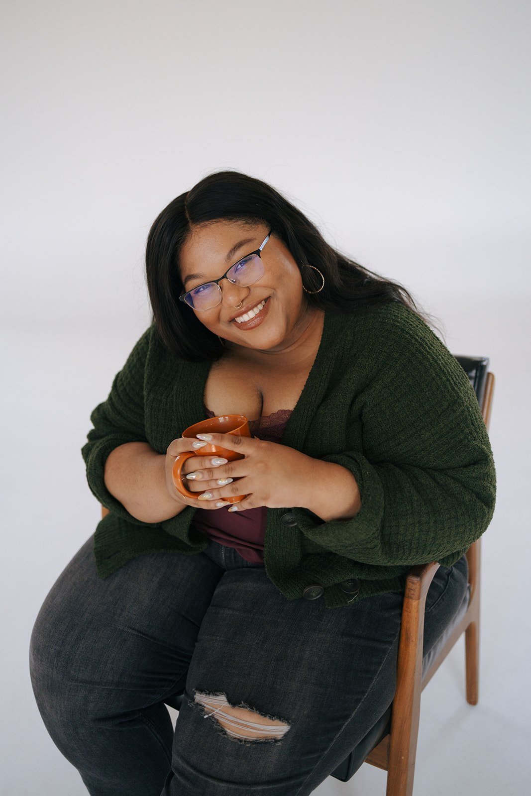 A smiling woman with glasses, hoop earrings, and a nose ring, sitting on a wooden chair, holding an orange mug, wearing a dark green cardigan, burgundy top, and ripped black jeans against a plain white background.
