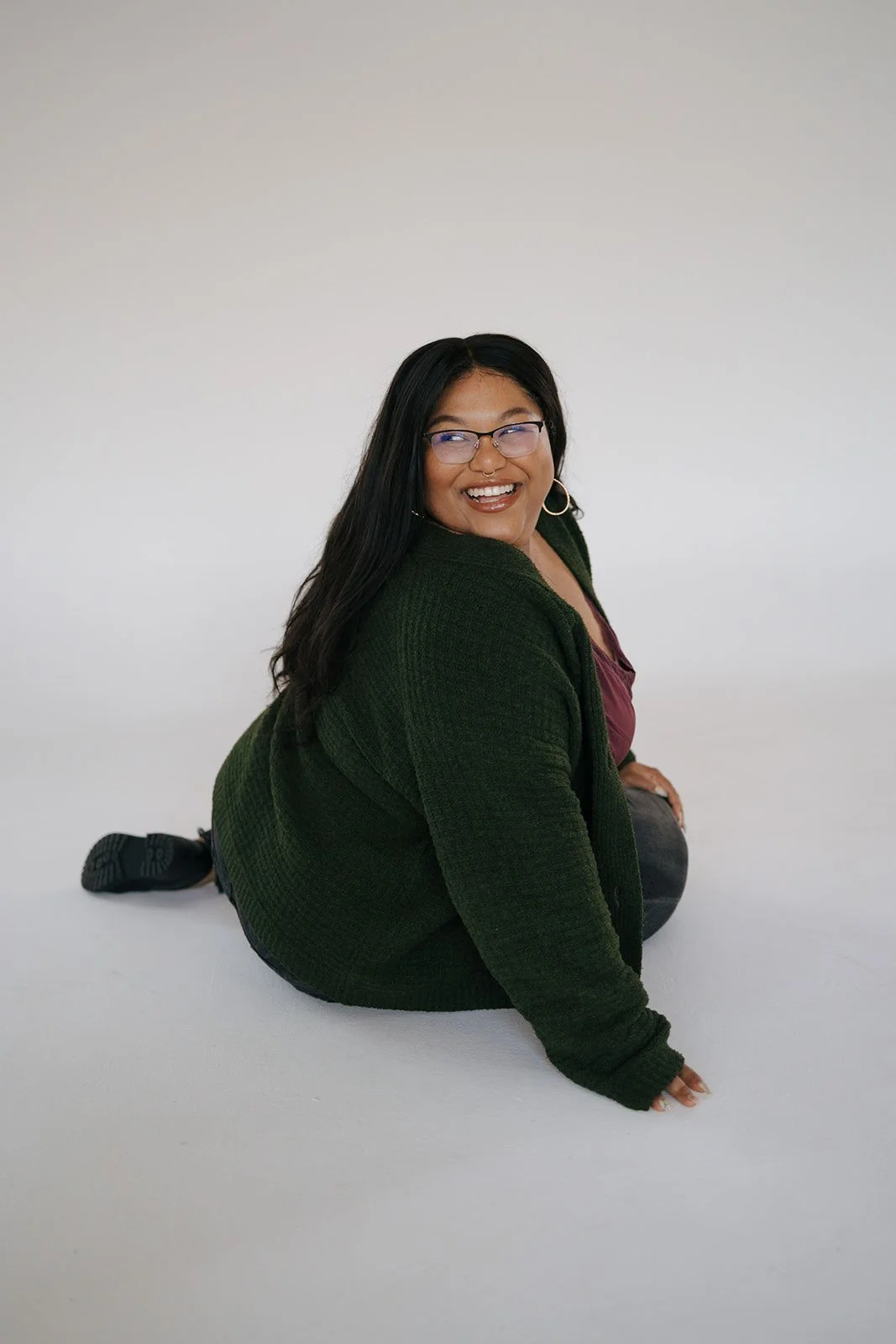 A smiling woman with long dark hair, wearing glasses, a green sweater, and dark pants, sitting on the floor against a plain white background.