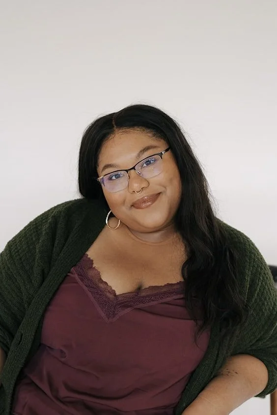 A woman with long dark hair, glasses, and a nose ring, smiling at the camera. She is wearing a dark green cardigan and a maroon top with lace detail.