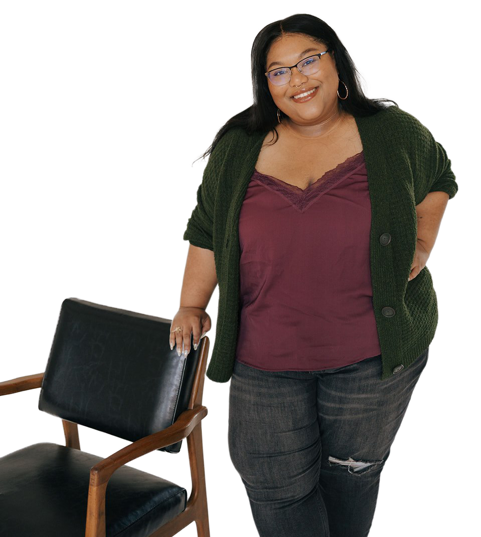 A smiling woman with glasses and hoop earrings standing next to a black chair with a wooden frame, wearing a purple top, green cardigan, and gray jeans with a tear on one knee.