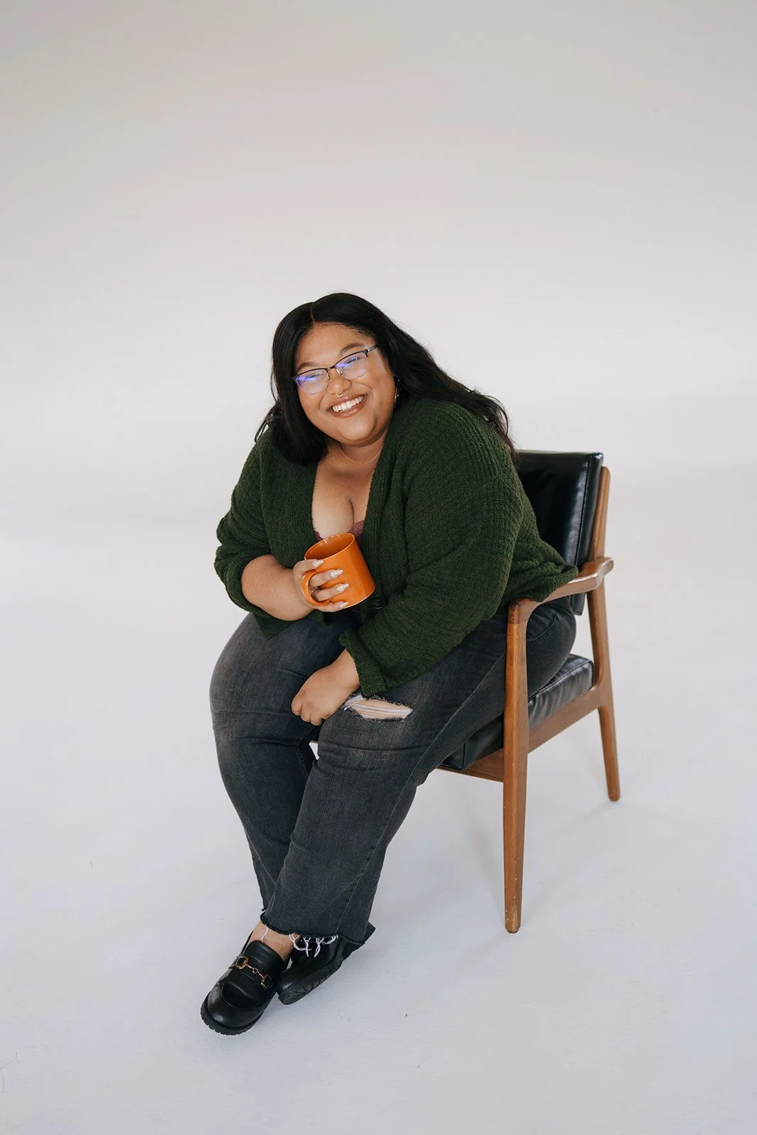 A woman with glasses, black hair, and a green sweater sitting on a wooden chair, smiling and holding an orange mug.