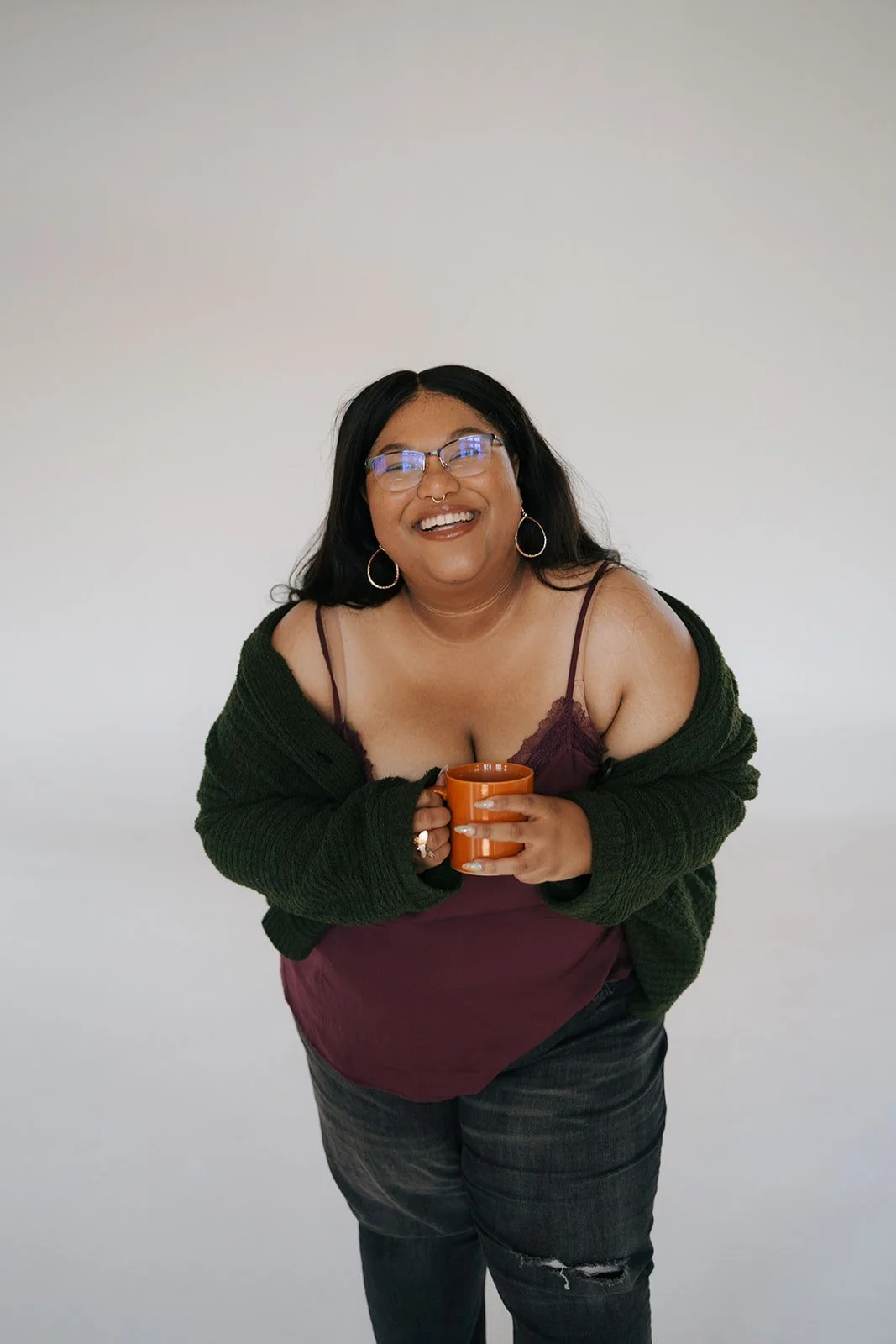 A smiling woman with black hair, glasses, and hoop earrings holding an orange mug, wearing a maroon camisole, and a dark green cardigan against a plain white background.