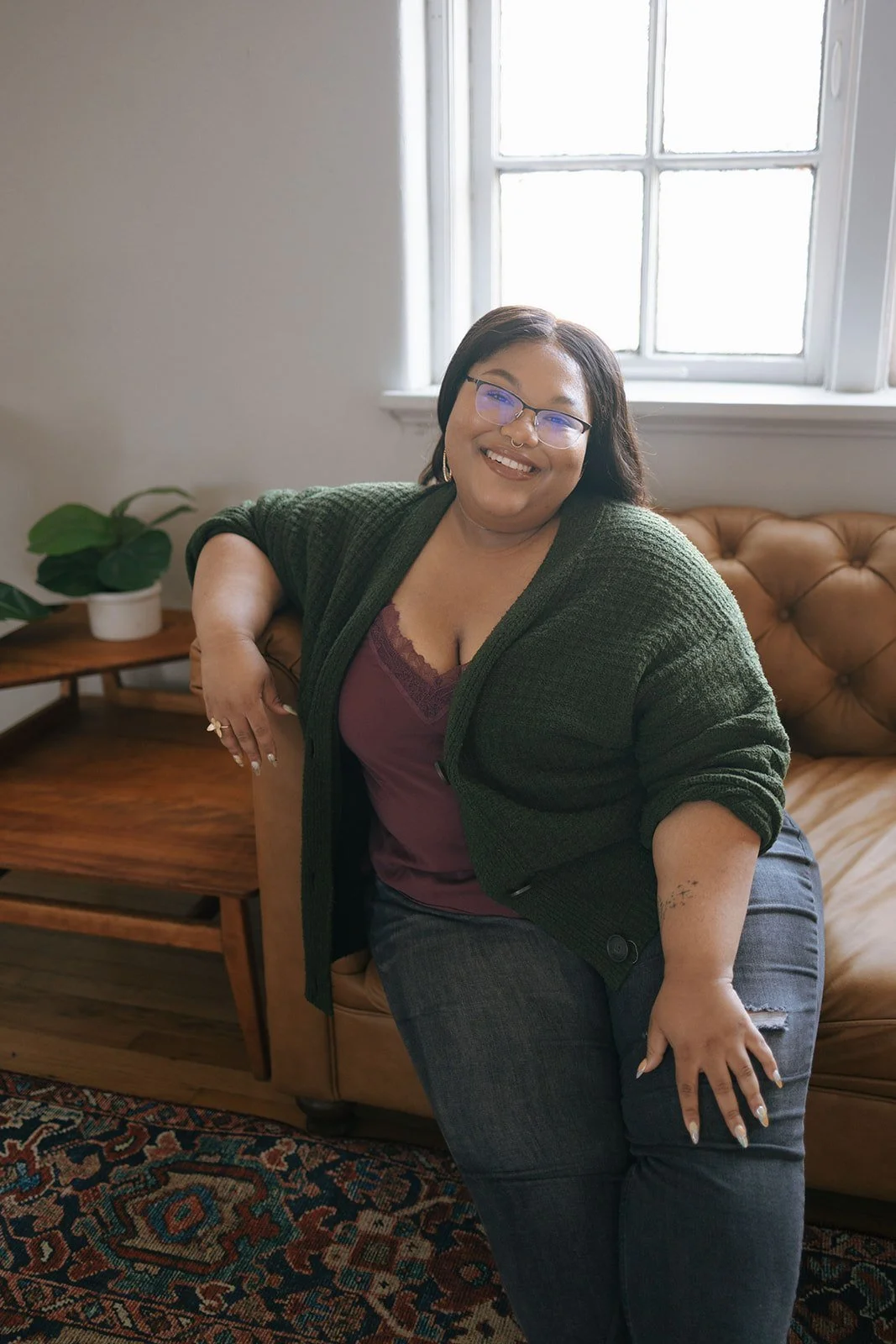 A woman sitting on a leather couch near a window with white framing, smiling and wearing glasses, a green sweater, and a maroon top, with a potted plant on a side table nearby.