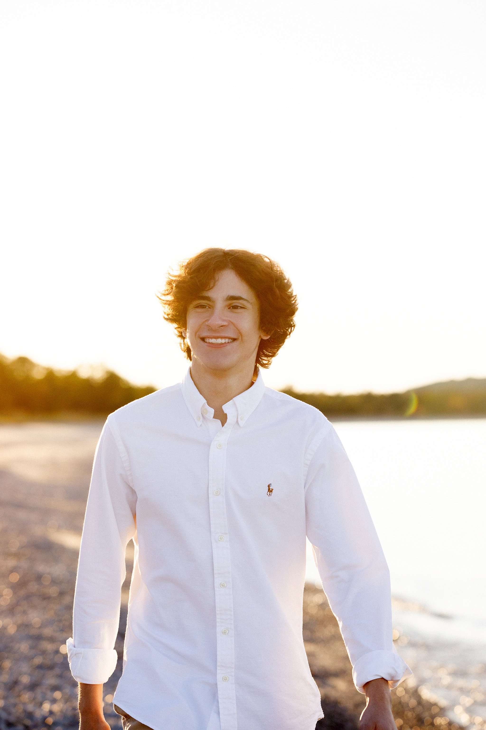 A young man with curly hair and a white button-up shirt smiling and walking on a beach at sunset.