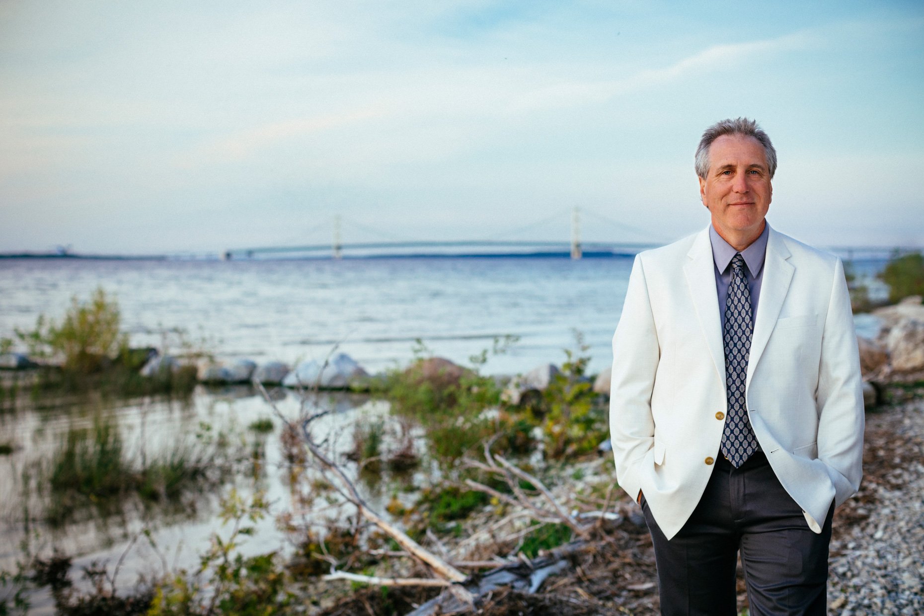 A middle-aged man in a white blazer and patterned tie standing on a rocky shoreline with water and a bridge in the background.