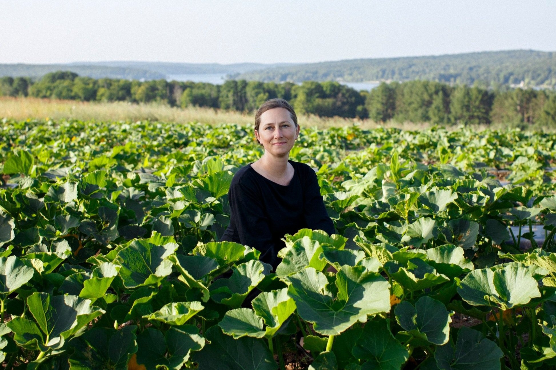 Woman in black shirt sitting in a lush green field of plants with a lake and distant hills in the background.