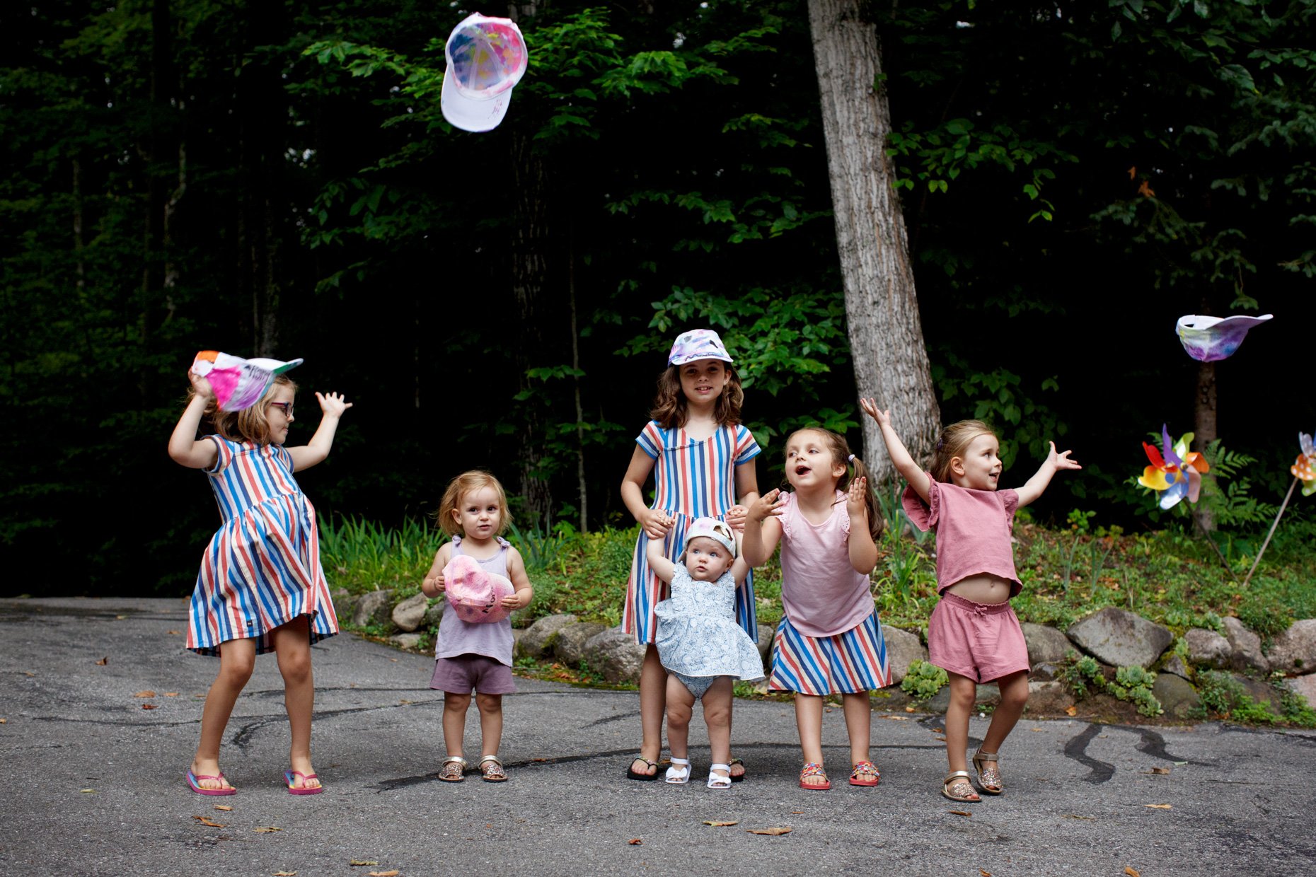 Group of six young girls celebrating outdoors with hats flying in the air and trees in the background.