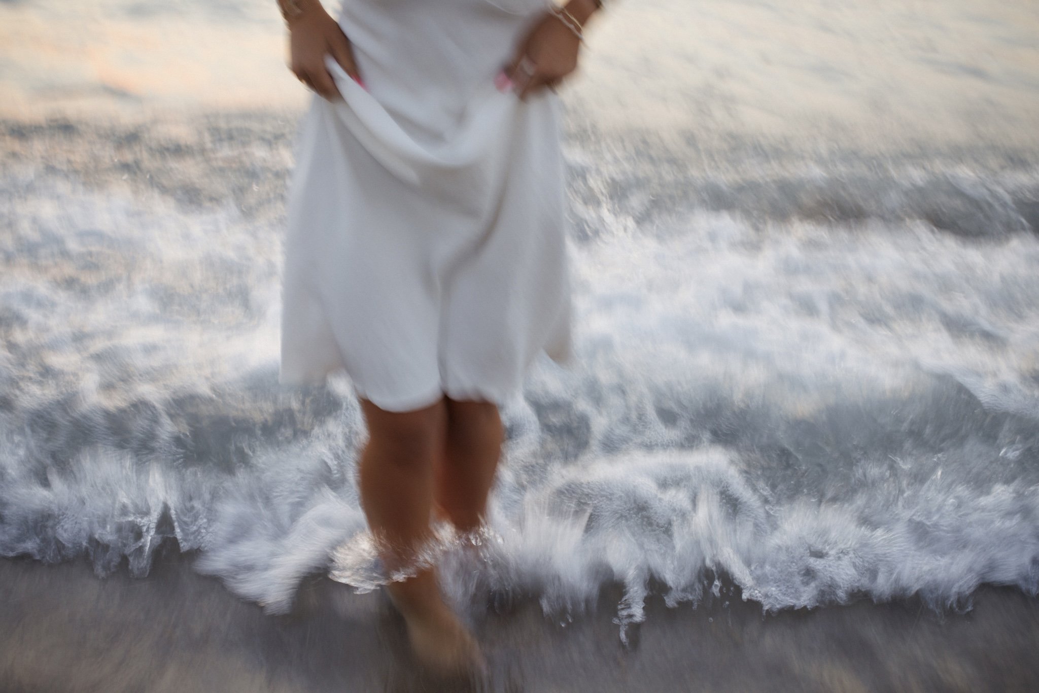 Person wearing a white dress walking in the shallow surf at the beach during sunset.