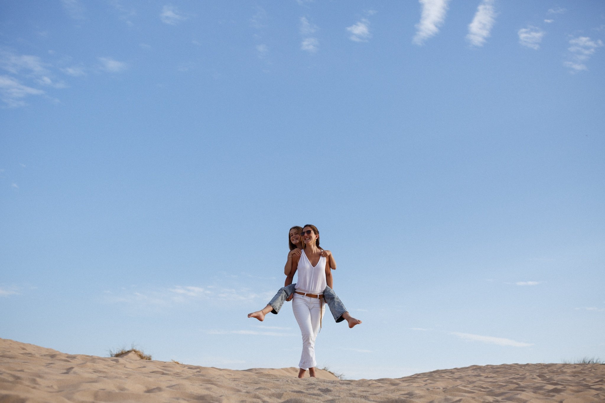 Two women smiling and enjoying each other's company at the beach, with one giving the other a piggyback ride on the sandy shore under a blue sky with a few clouds.