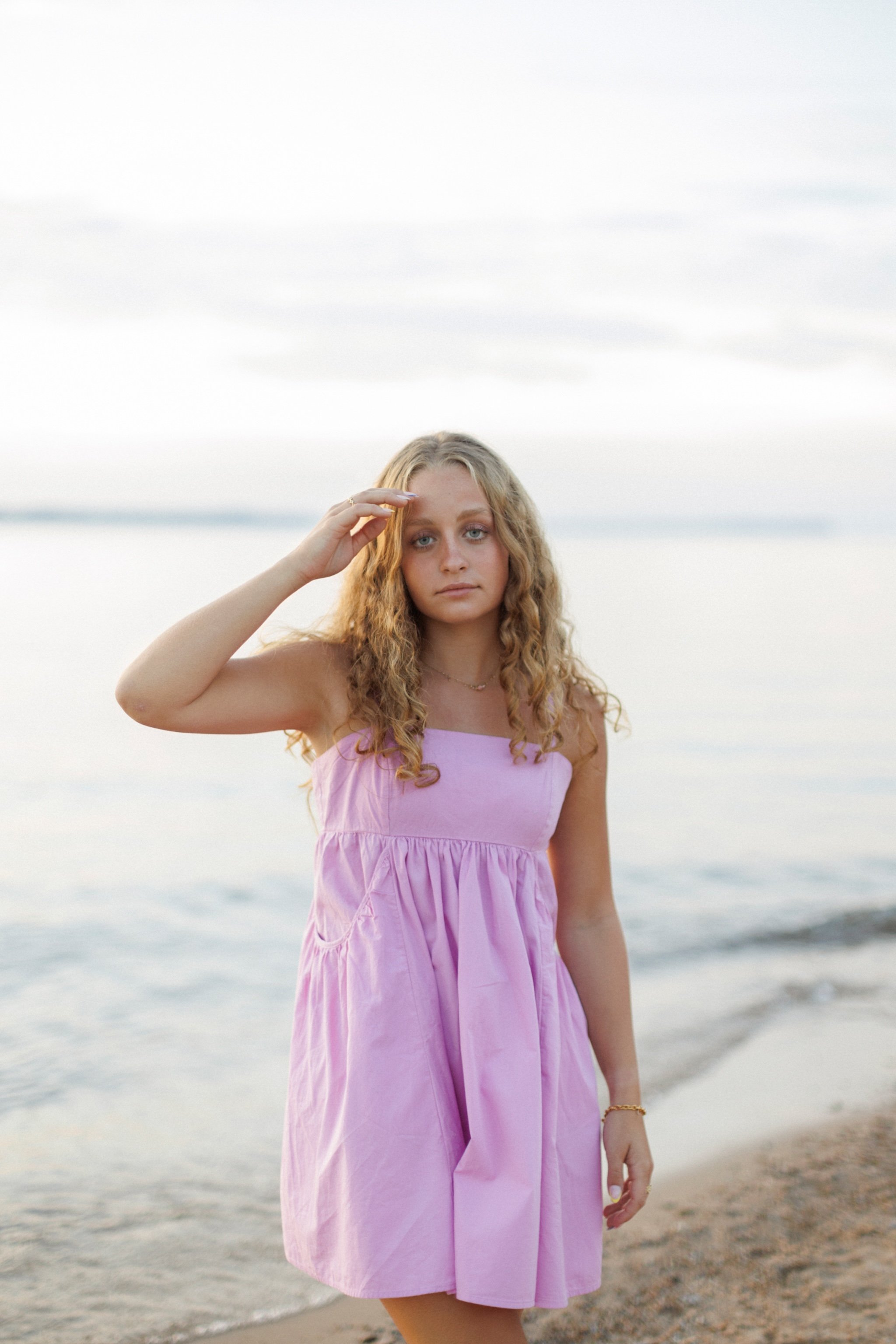 A young girl with curly blonde hair gazes at the camera while standing on a beach near the water, wearing a pink dress, with her hand shielding her eyes.