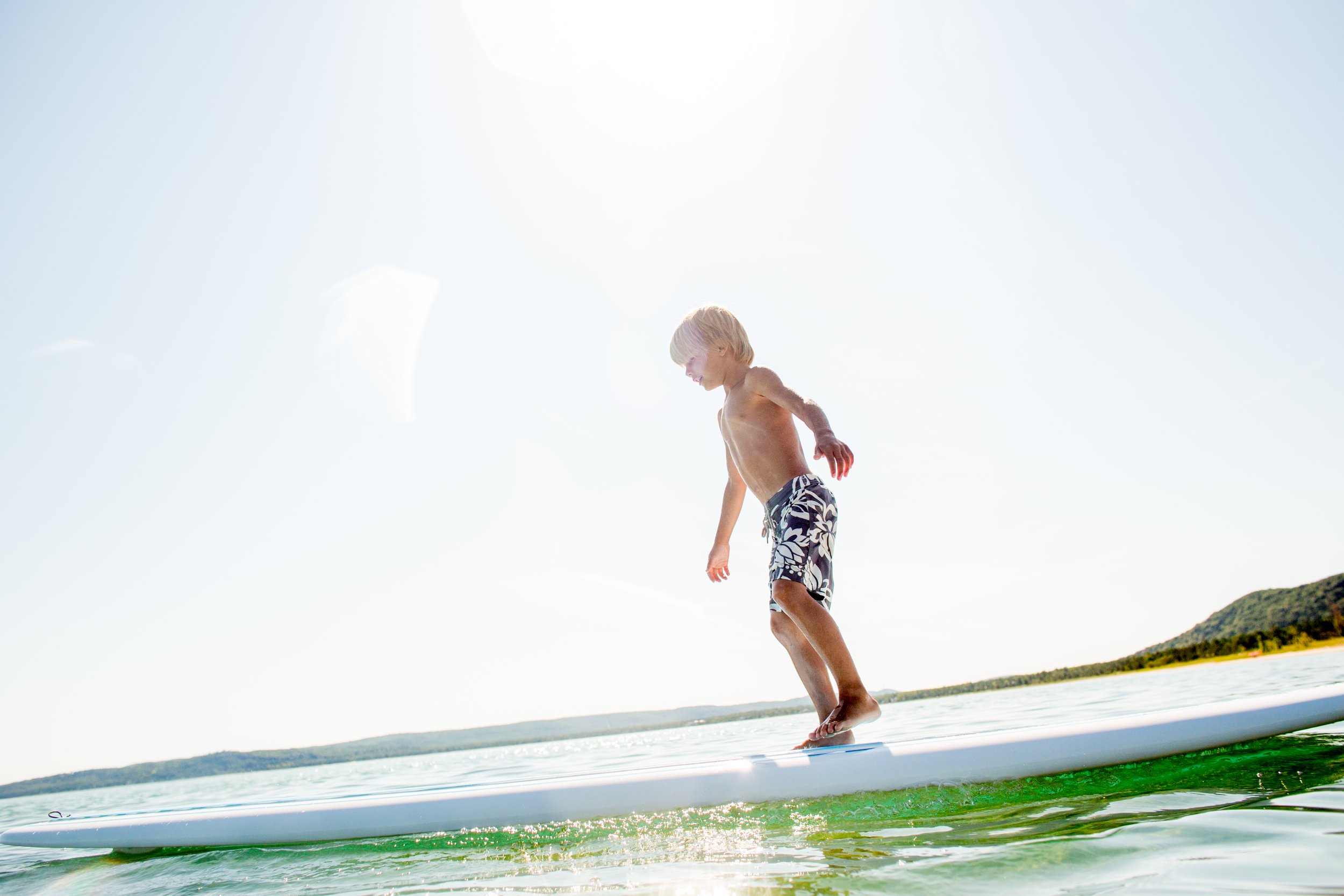 A young boy with blonde hair is standing barefoot on a white paddleboard floating on water, wearing swim trunks with a floral pattern, under a bright sun in a clear sky.