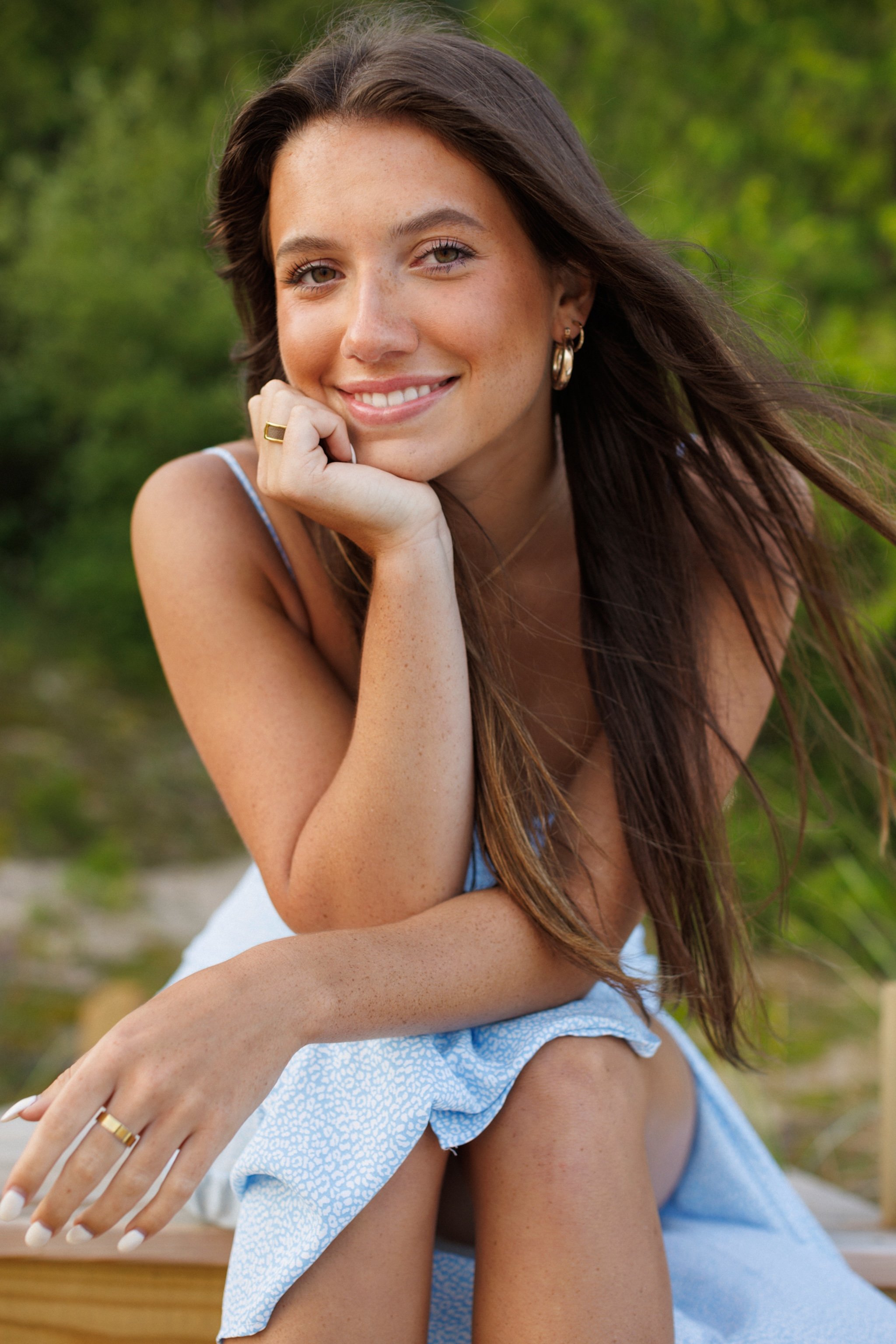 A young woman with long brown hair, wearing a light blue dress, smiling and resting her chin on her hand outdoors with green foliage in the background.