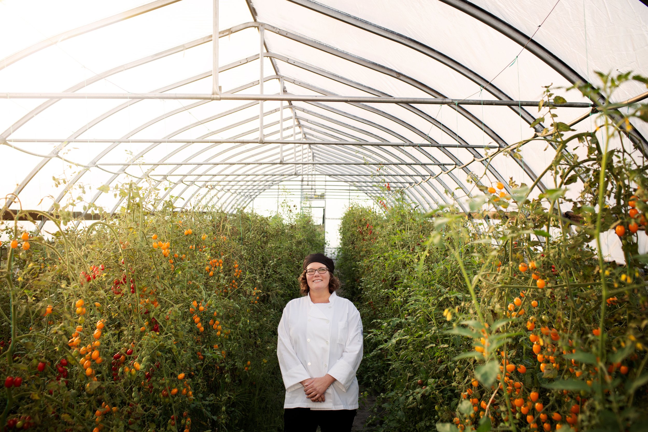 A woman in a white chef's coat and glasses stands in a greenhouse filled with tomato plants bearing red and orange cherry tomatoes.