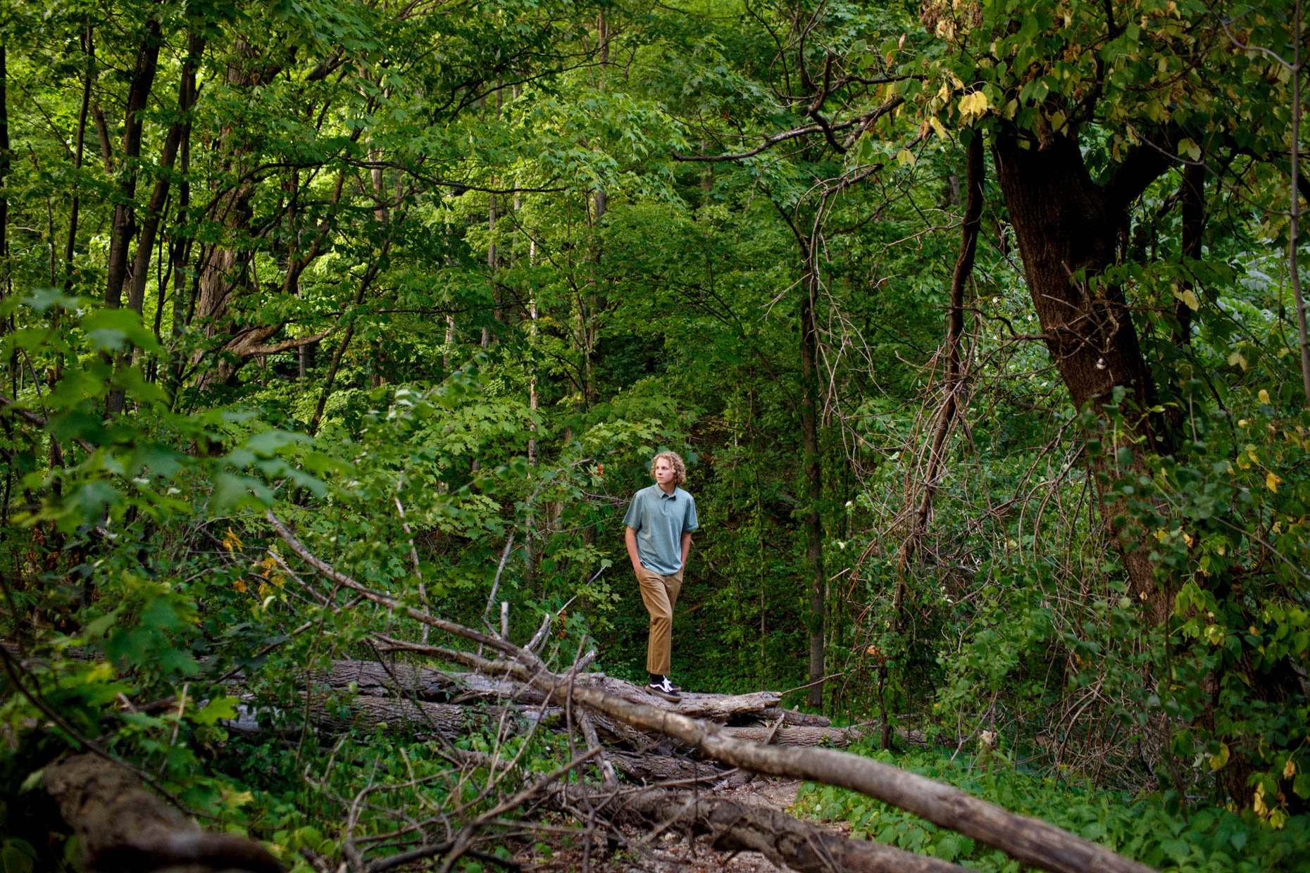A young man with curly blonde hair wearing a green shirt, khaki pants, and sneakers stands on a fallen tree trunk in a dense green forest, looking to the side.