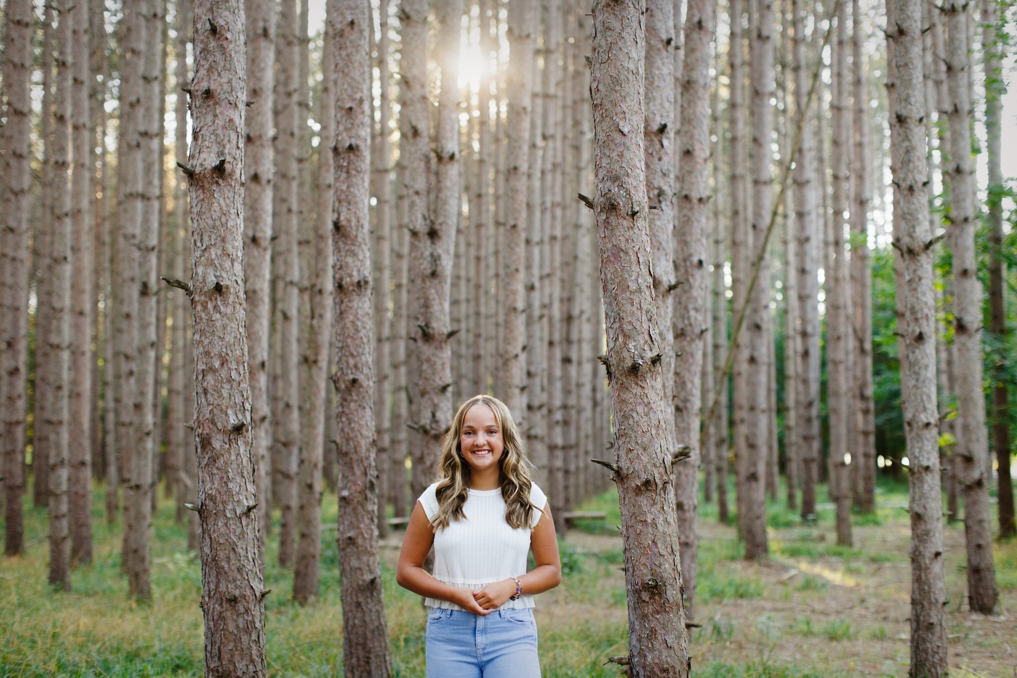 A young woman smiling in a forest of tall, thin pine trees, with sunlight shining through the trees.