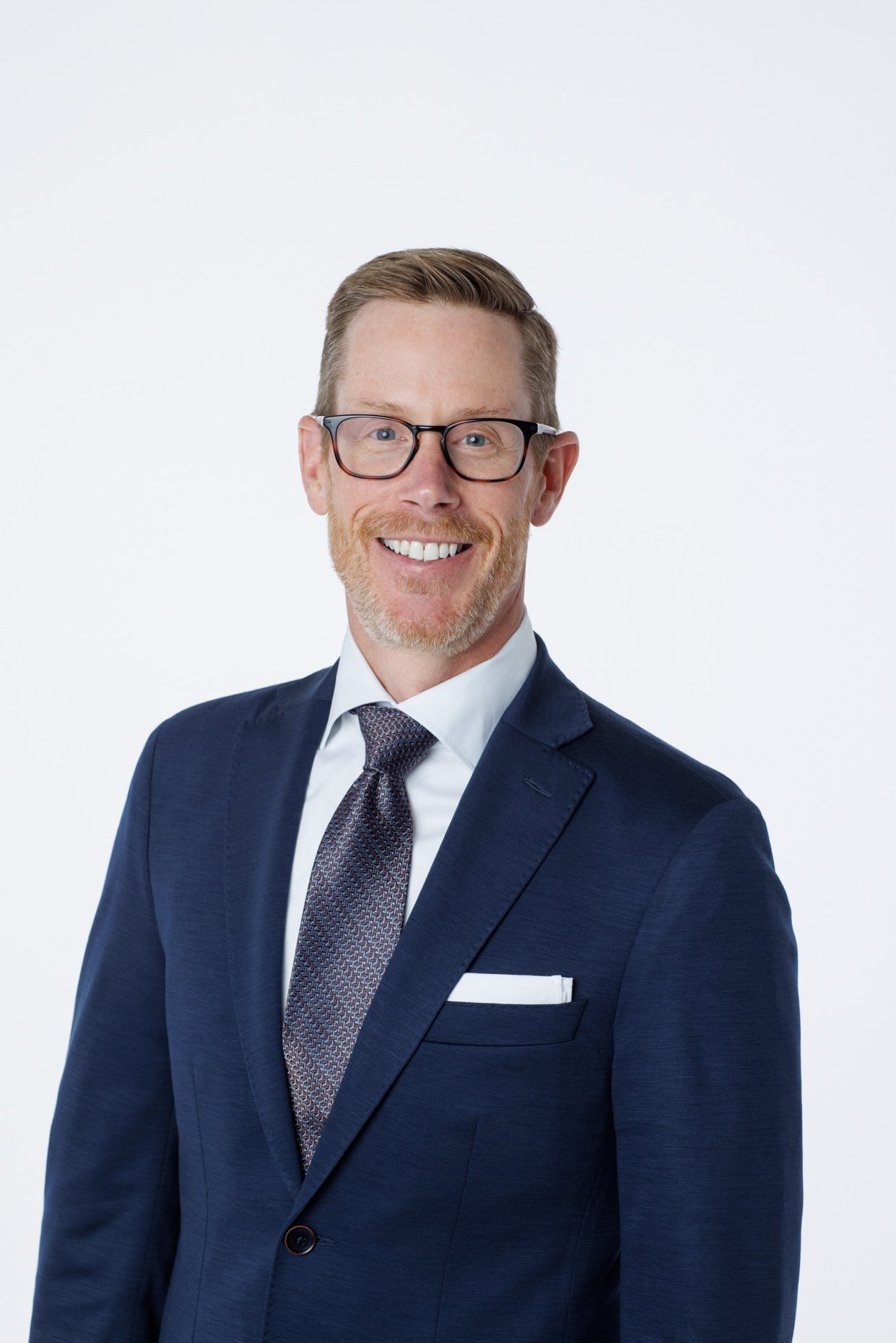 A professional man smiling in a navy suit, white shirt, patterned tie, and glasses against a plain white background.