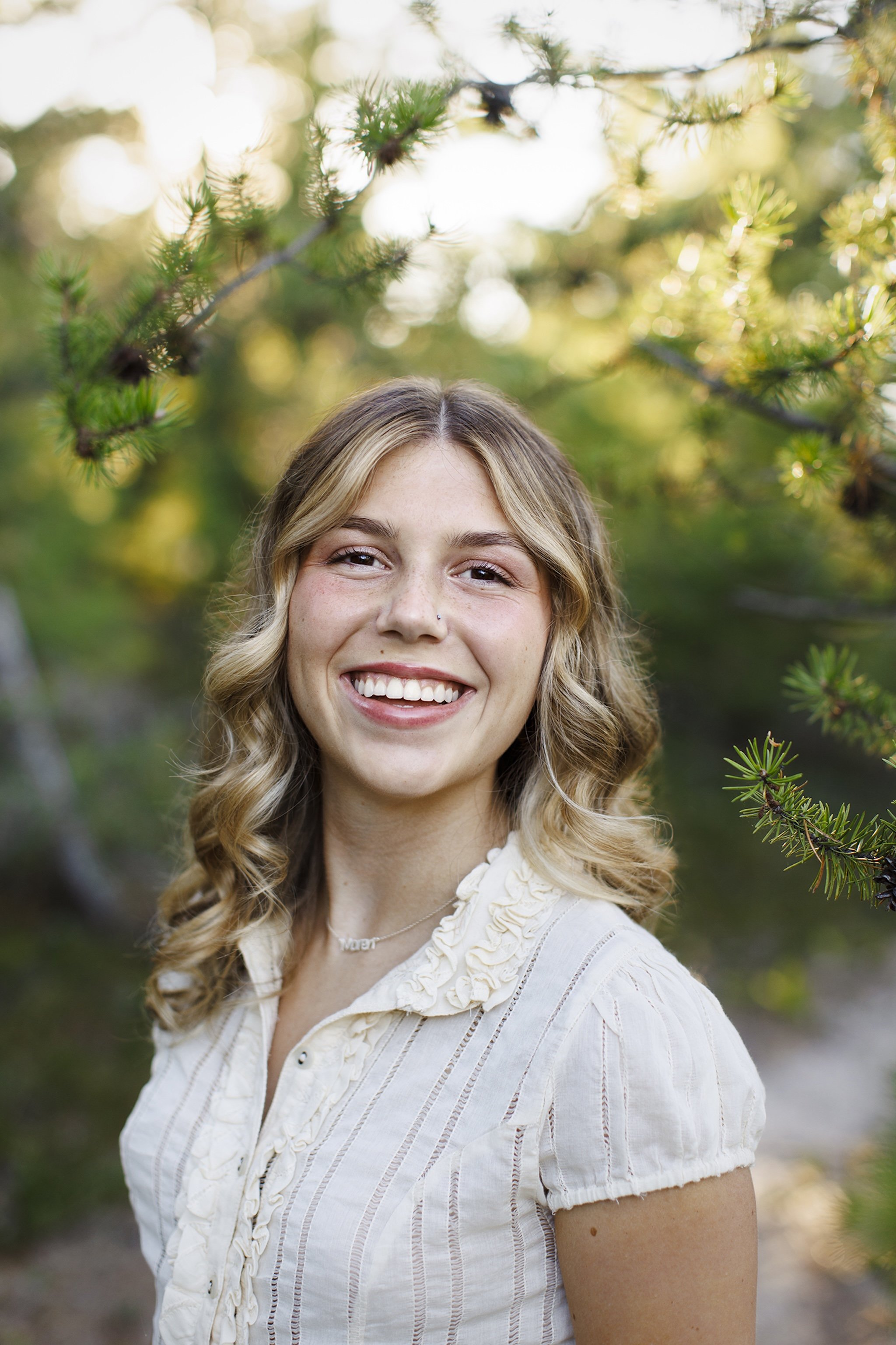 A smiling young woman with blonde, curly hair outdoors among green trees.