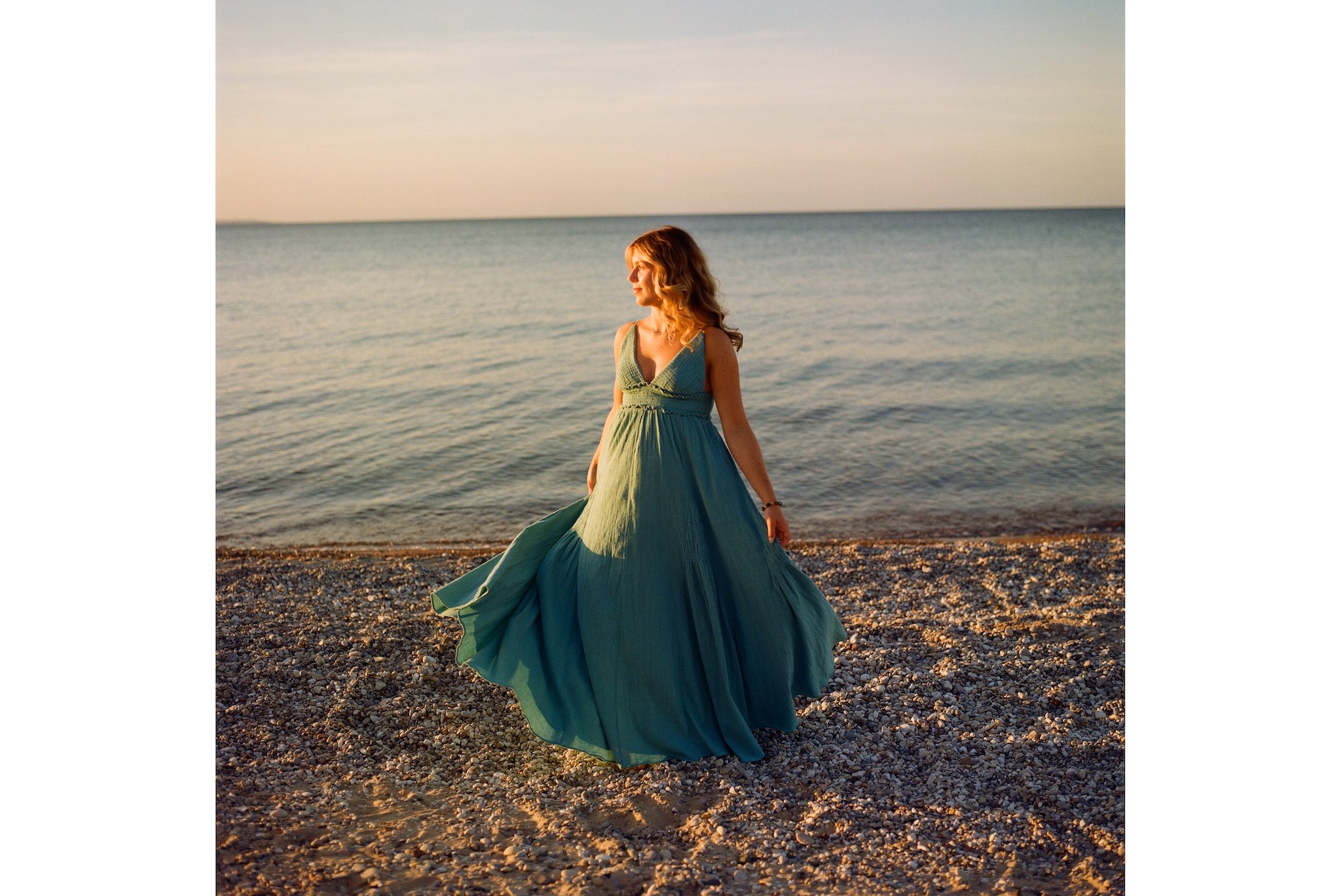 A woman in a flowing, teal dress standing on a pebbled beach during sunset, facing slightly to the left with her hand resting on her dress.