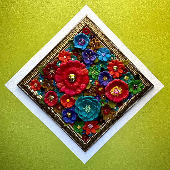 Colorful felt flowers with beads and decorative elements arranged in a diamond shape in a gold frame on a yellow wall.
