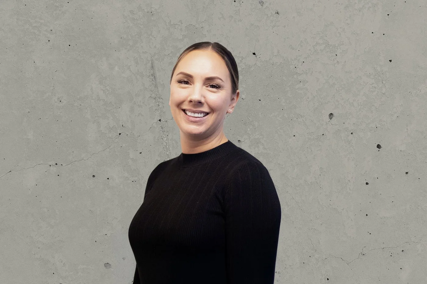 A smiling woman with light skin and dark hair tied back, wearing a black long-sleeve shirt, standing against a textured gray concrete wall.