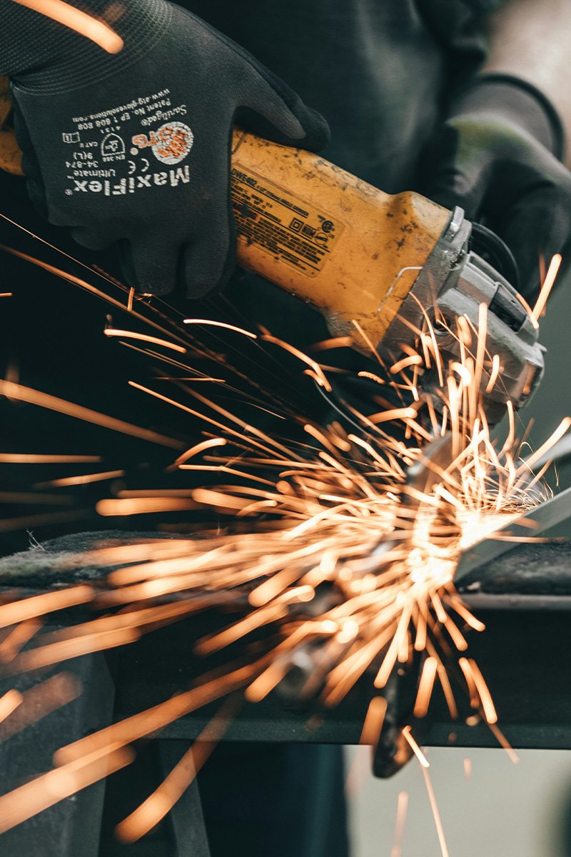 A person wearing black gloves is using an orange power angle grinder to cut metal, producing sparks.