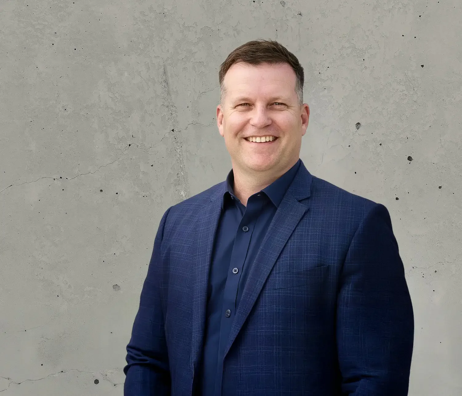 Richard Keown wearing in a blue suit and navy shirt standing against a textured concrete wall.