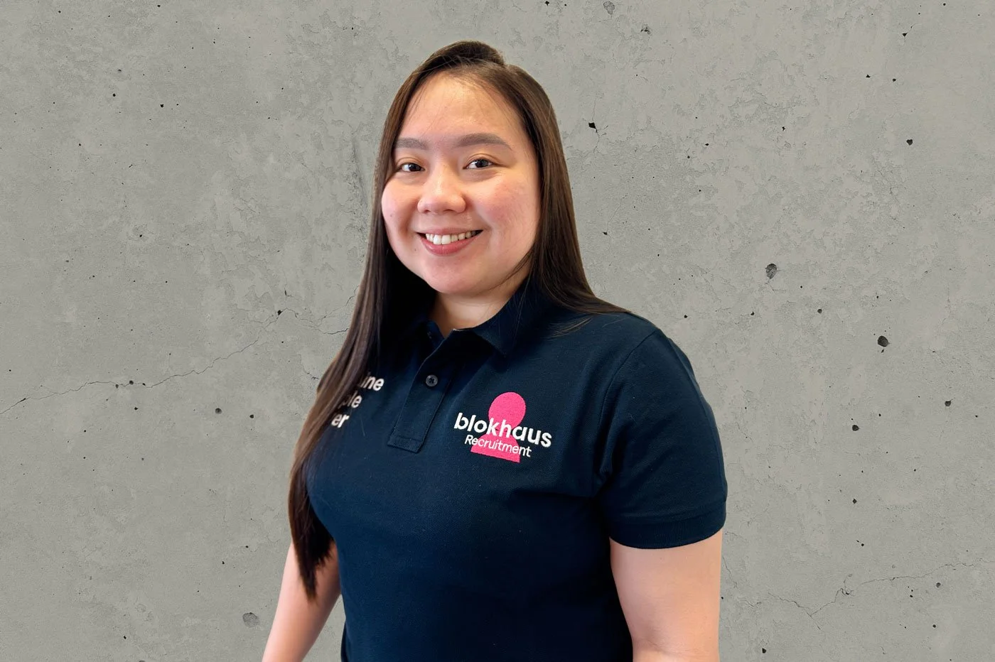 A woman with long dark hair smiling at the camera, wearing a navy blue polo shirt with the Blokhaus Recruitment logo, standing against a gray concrete wall.