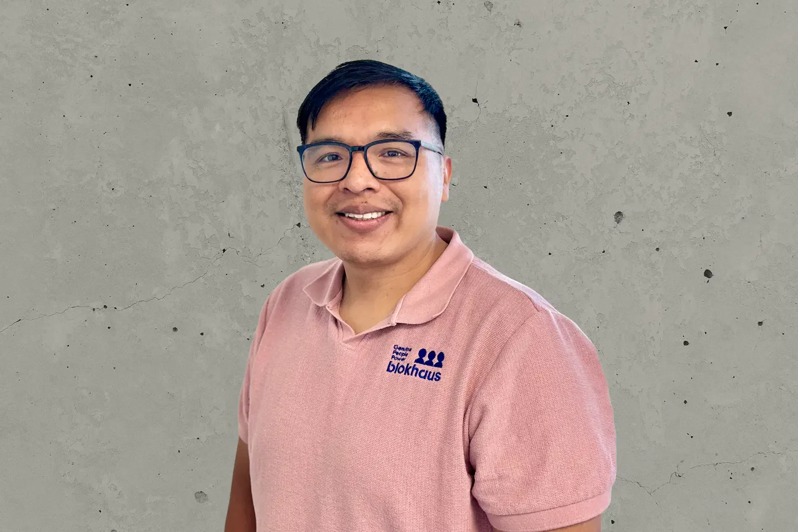 A smiling man wearing glasses and a blush pink polo shirt with 'Blokhaus' logo standing against a textured gray wall.