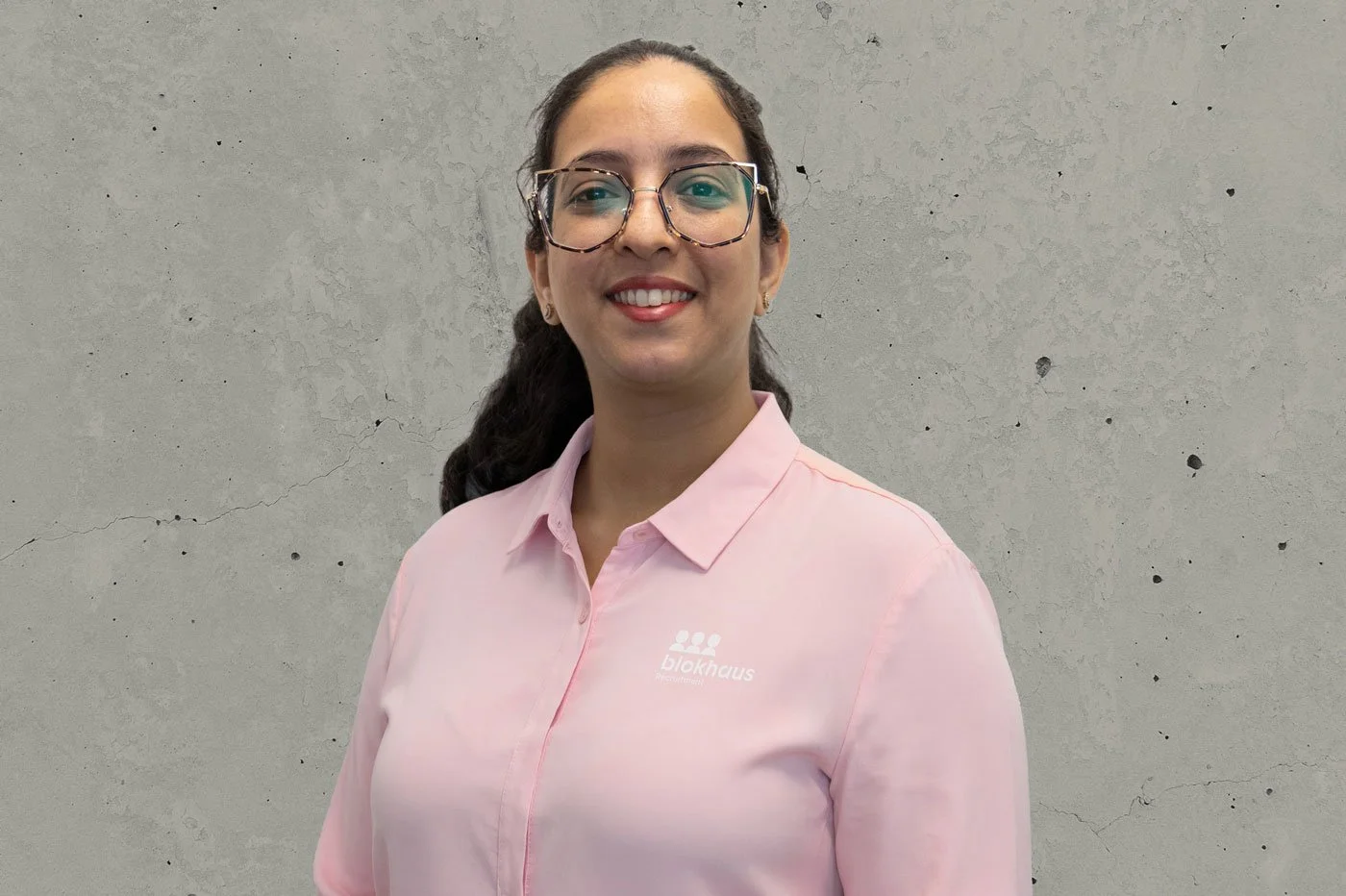 A woman with glasses and long dark hair wearing a light pink button-up shirt with a logo on the chest, standing against a gray wall with cracks.