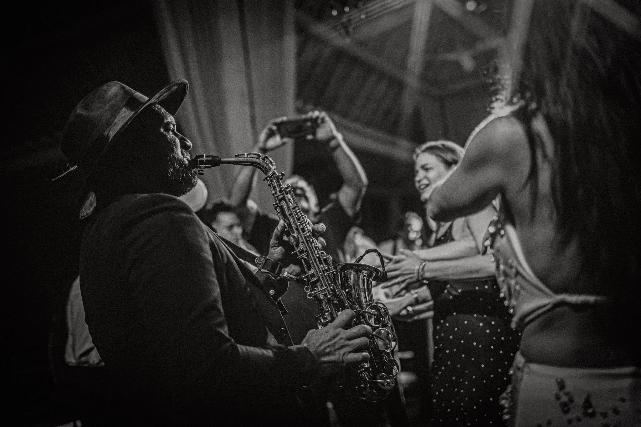 Hombre tocando saxofón en un concierto con varias personas bailando y disfrutando de la música en un ambiente oscuro.