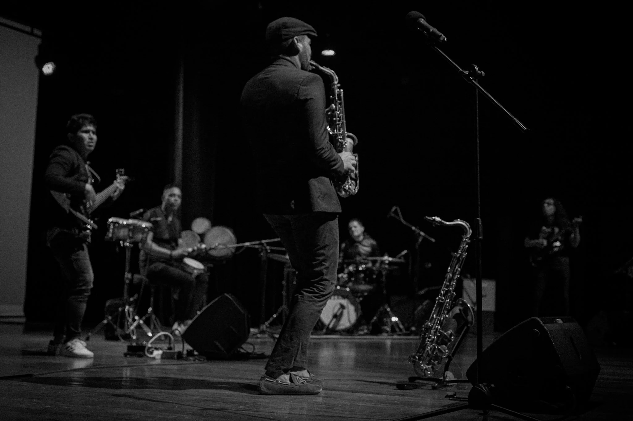 Un grupo de músicos toca en un escenario oscuro, con un saxofonista en primer plano y otros tocando instrumentos como guitarra, batería y percusión.