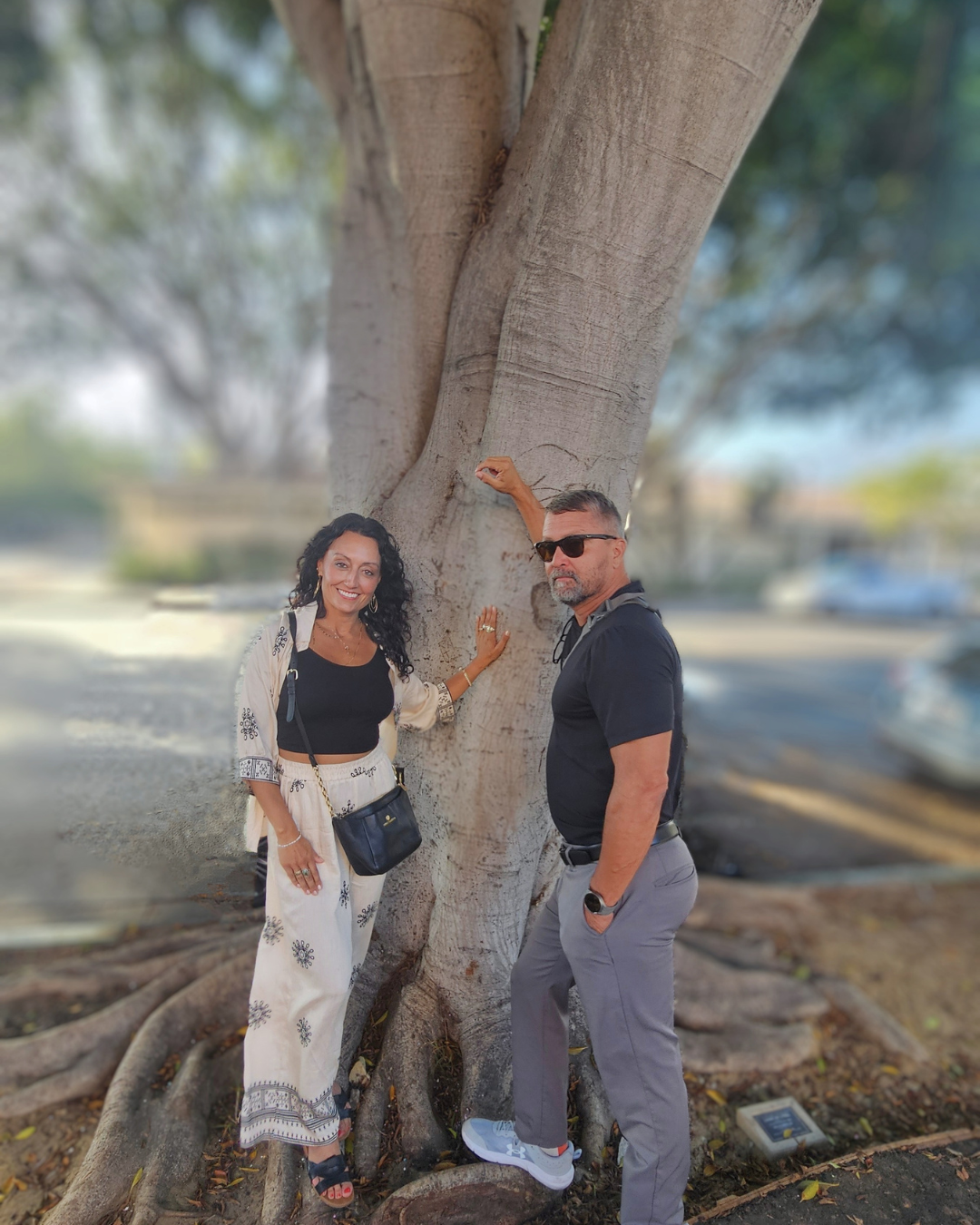 A man and a woman standing next to a large tree with wide roots, outdoors, in a park or parking lot, during daytime. The woman has long, curly dark hair, and the man has short gray hair and is wearing sunglasses.