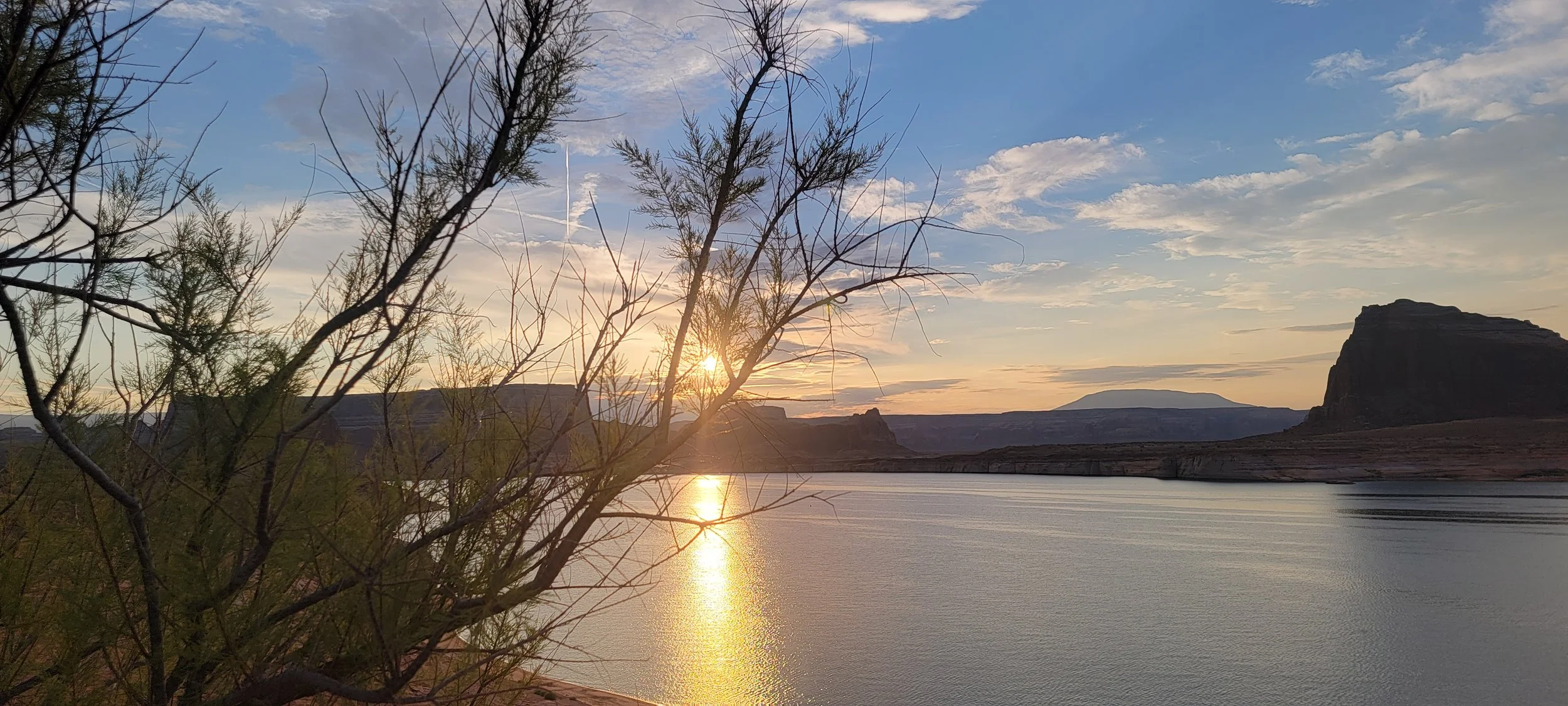 Scenic landscape of a river at sunset with rocky mesas, a tree in the foreground, and a partly cloudy sky.
