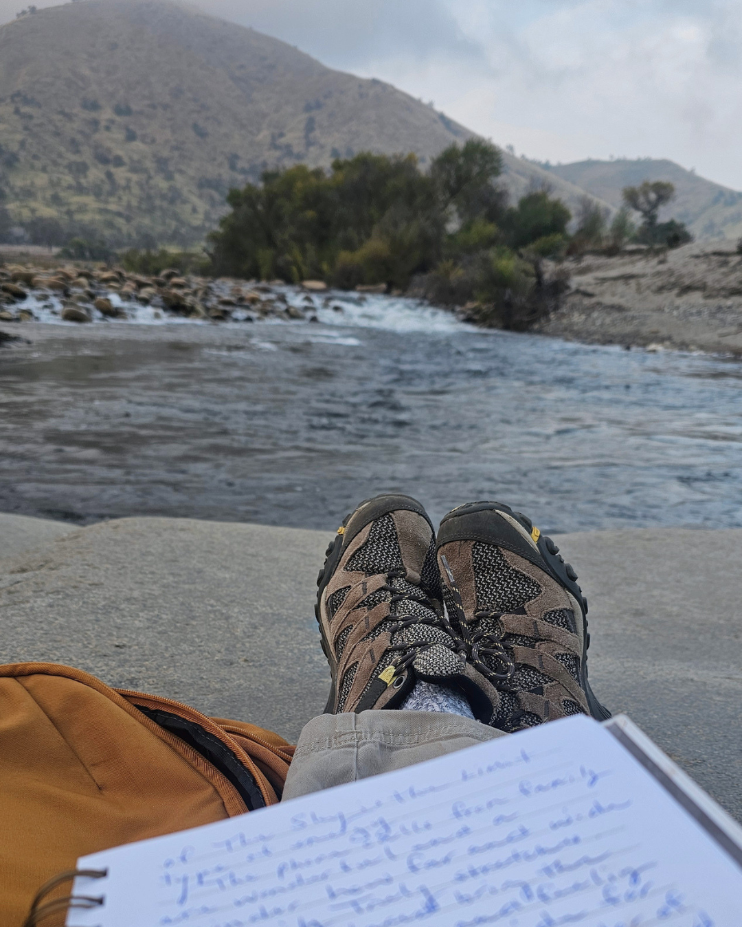 Person sitting near a river, wearing hiking boots, with a notebook and a backpack visible in the foreground, and mountains, trees, and flowing water in the background.