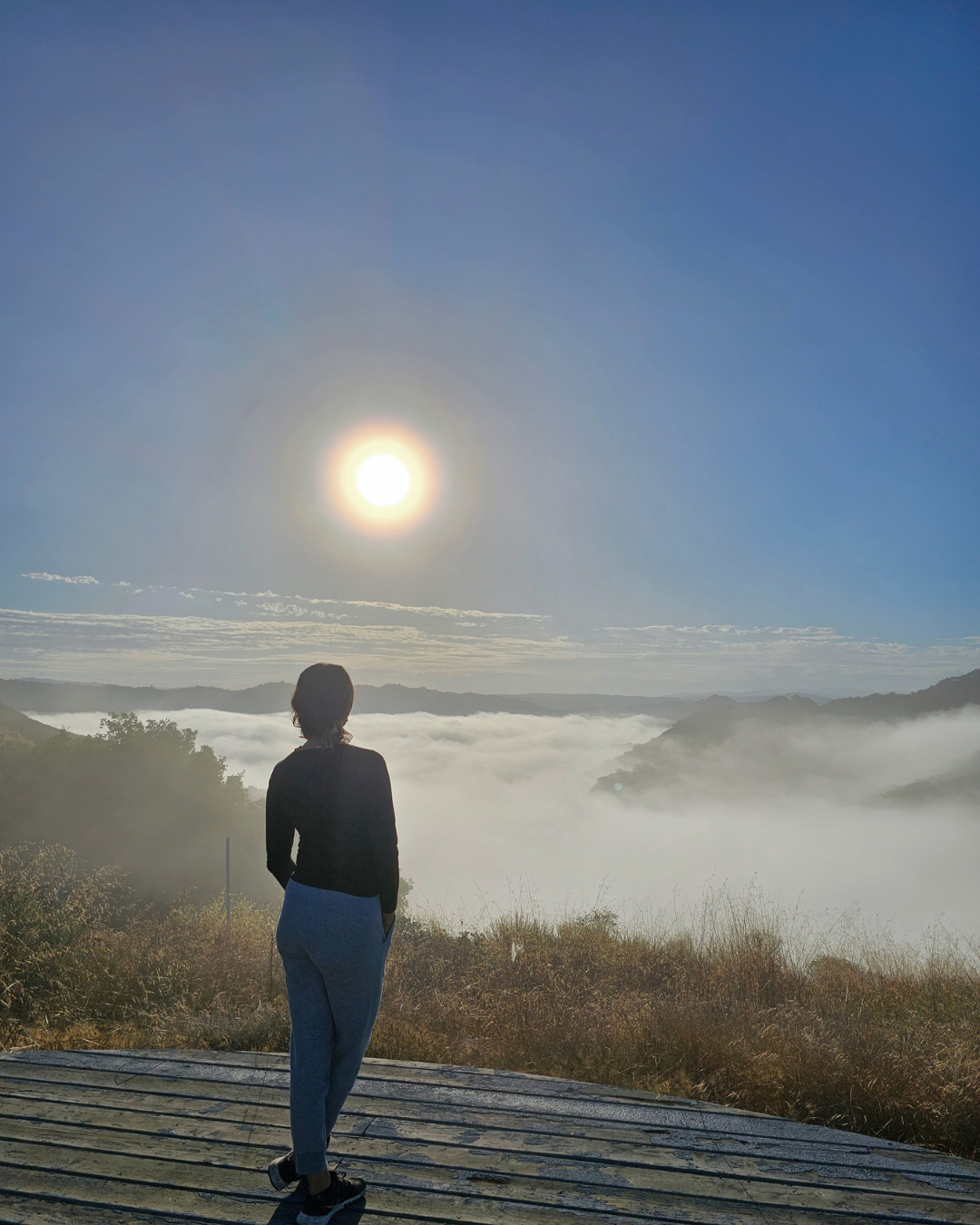Person standing on a wooden platform overlooking a foggy valley with hills, under a clear sky with the sun shining brightly.