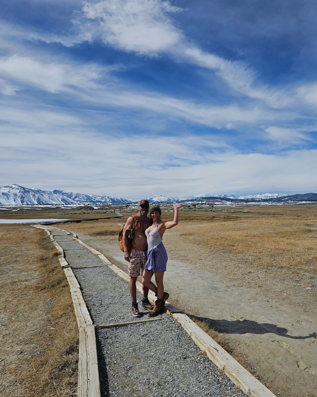 Two people standing on a gravel path in a flat open landscape with snow-capped mountains in the background under a partly cloudy sky.