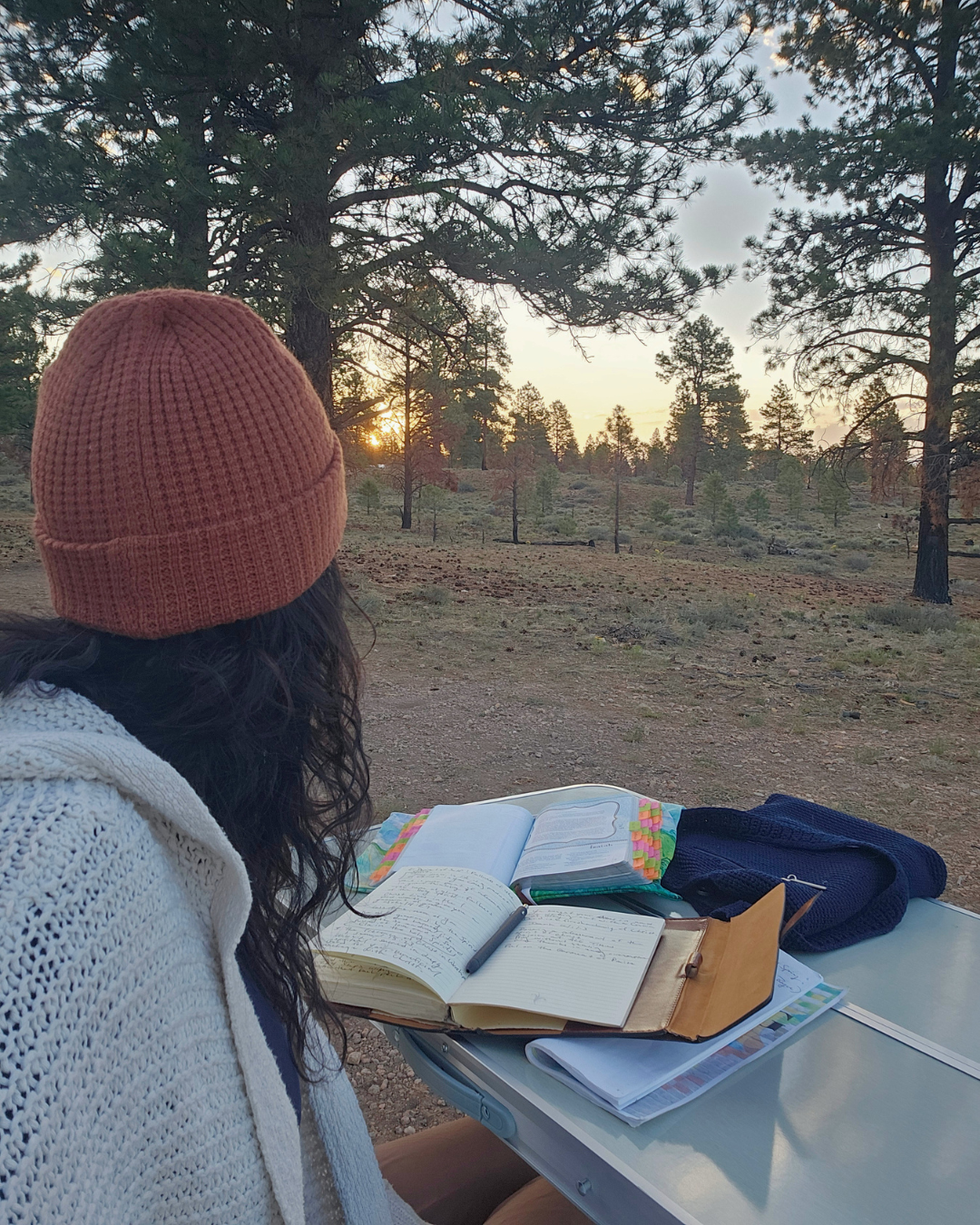 A woman with dark curly hair wearing an orange knit beanie and white sweater sits at a table outdoors during sunset, with notebooks and a pen in front of her, in a forested area with pine trees.