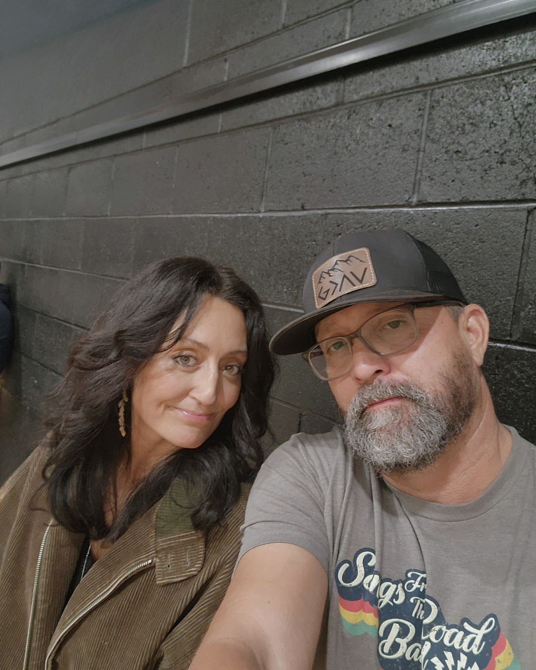A woman with dark wavy hair and a man with a beard, glasses, and a cap, taking a selfie against a black brick wall.