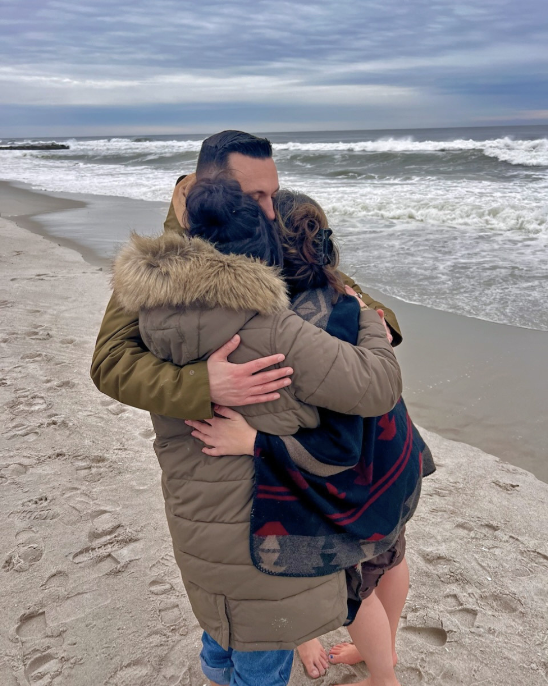 Three people embracing each other on a sandy beach with ocean waves and a cloudy sky in the background.