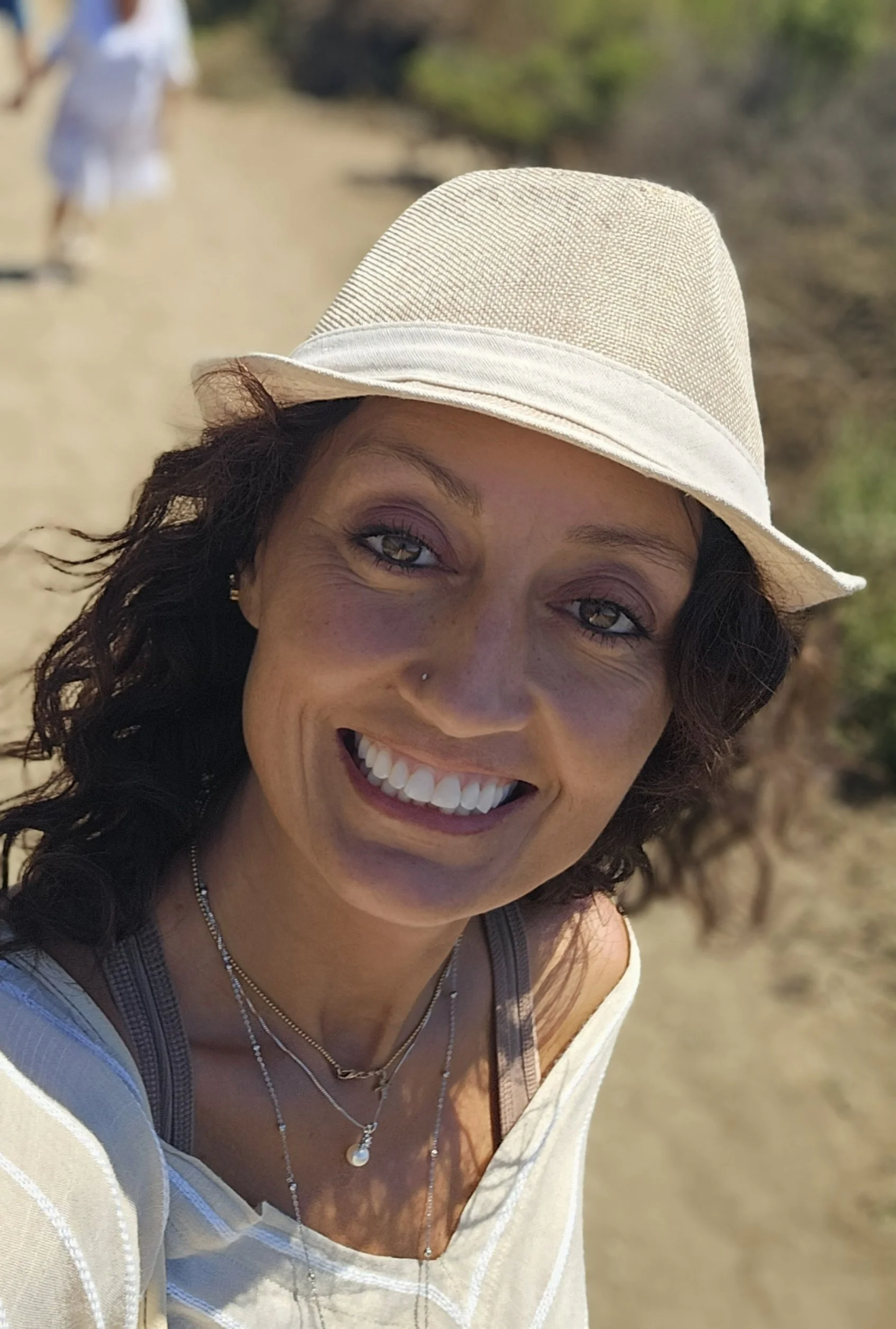 A woman smiling outdoors wearing a beige hat, layered necklaces, and a light-colored top, with a blurred background of a sandy path and greenery.