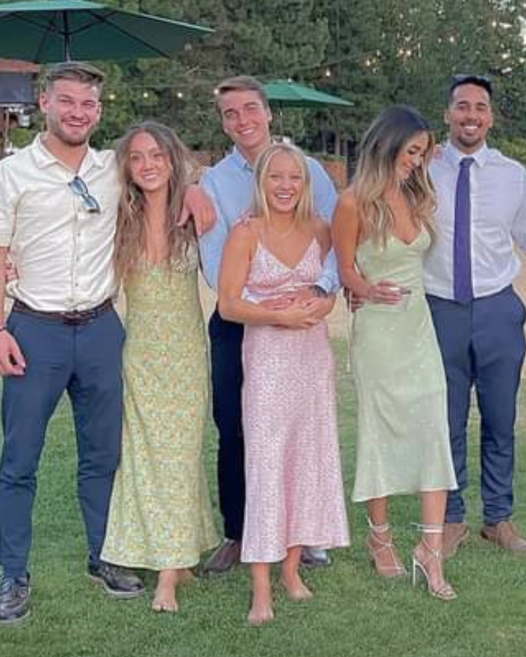 Group of six people dressed in semi-formal attire standing outdoors on grass, smiling and posing. Two men and four women, some holding glasses, with trees and string lights in the background.
