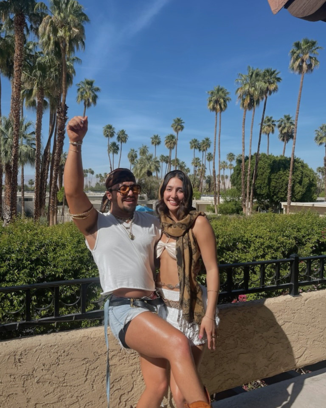 Two young women enjoying sunny weather outdoors with palm trees in the background. One woman is wearing a white crop top, denim shorts, sunglasses, and a headband, with her arm raised. The other woman is wearing a brown scarf, white top, and a skirt. They are smiling, standing next to a low wall with green bushes behind them.