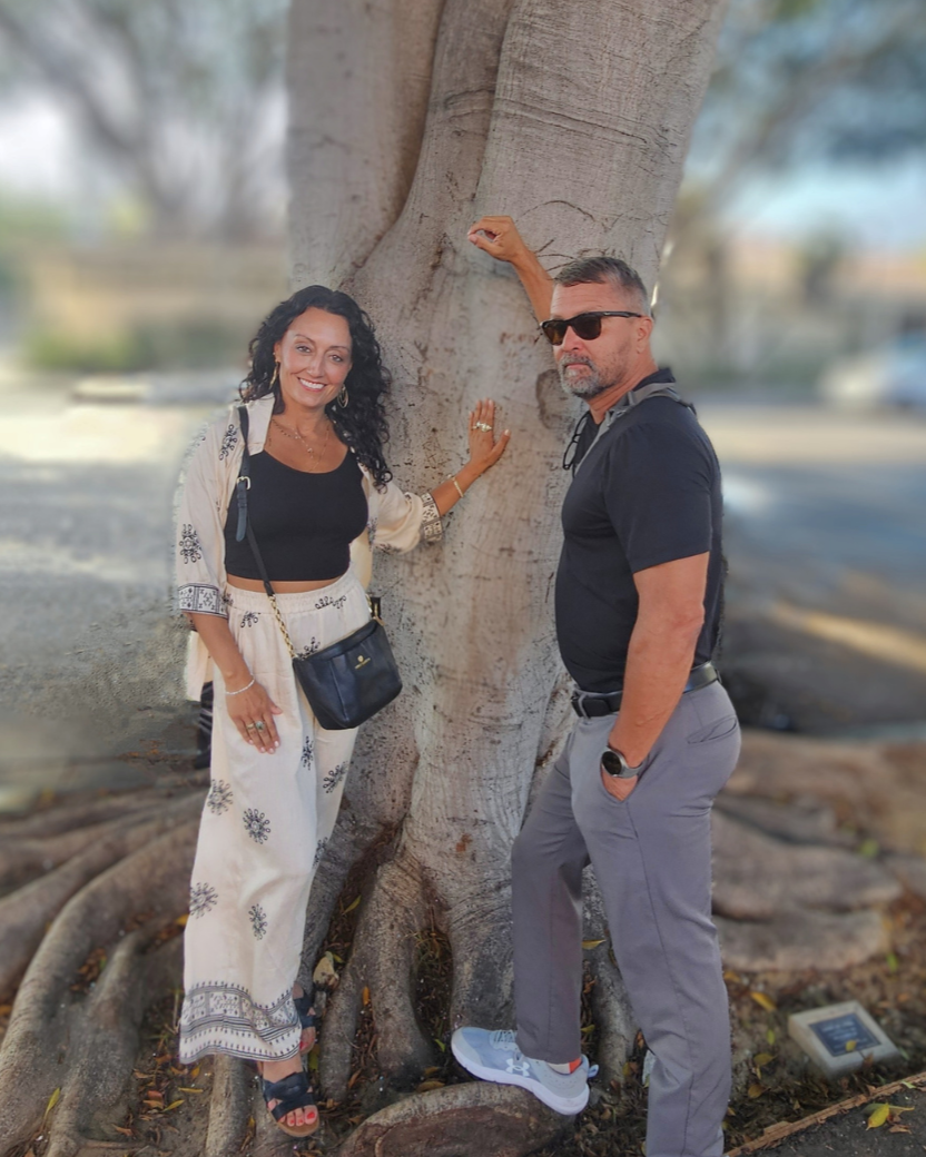 A woman with dark curly hair wearing a white embroidered outfit and a man with short hair and sunglasses wearing a black polo shirt and gray pants standing beside a large tree with visible roots, outdoors during daytime.